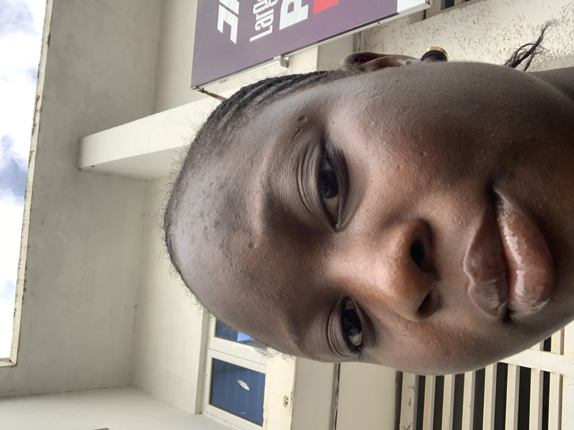 A close-up, low-angle selfie shows a young Black woman from the chest up. She has dark skin, braided hair, and is wearing a gold and dark hoop earring. Her lips are glossy. Behind her, a building with white walls and a visible roof structure is visible, along with a sign with purple and white lettering. The scene appears to be outdoors in an urban environment.