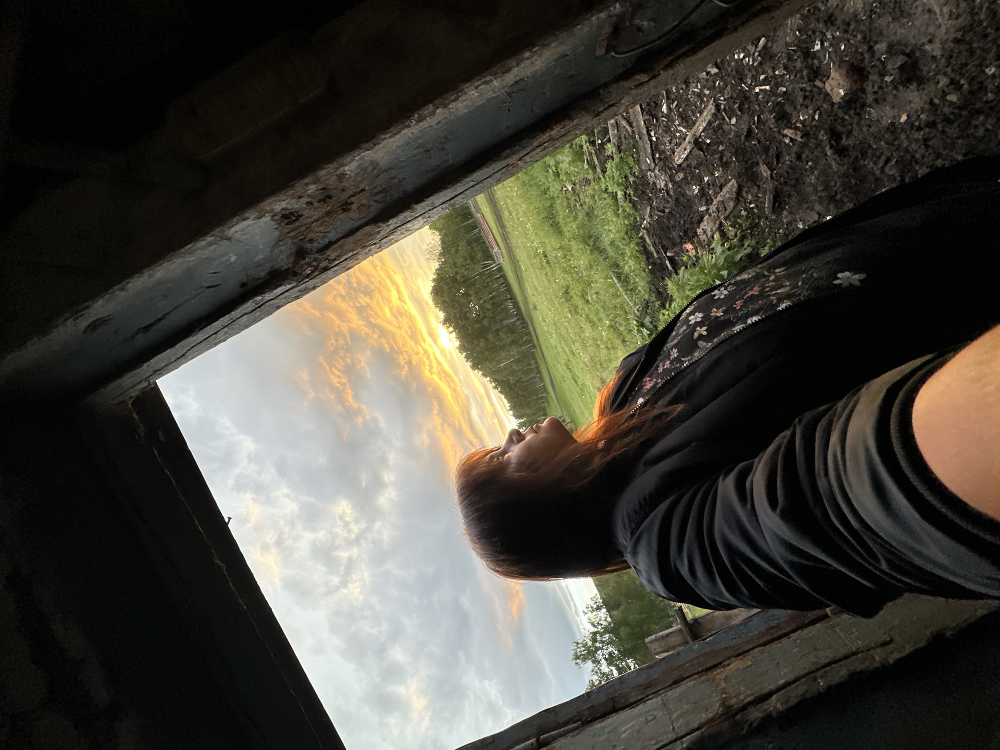 A woman with shoulder-length reddish-brown hair looks upwards and to the right, her face illuminated by the setting sun, which is visible in the distance beyond a green field. She is wearing a black jacket with a floral pattern on the inner lining. The image is framed by dark, weathered wooden structures, suggesting she is standing within an opening of some kind, possibly a doorway or window. The foreground is muddy and uneven, with scattered debris. The background features a line of trees against a sky filled with dramatic clouds, tinged with orange and yellow hues from the sunset.