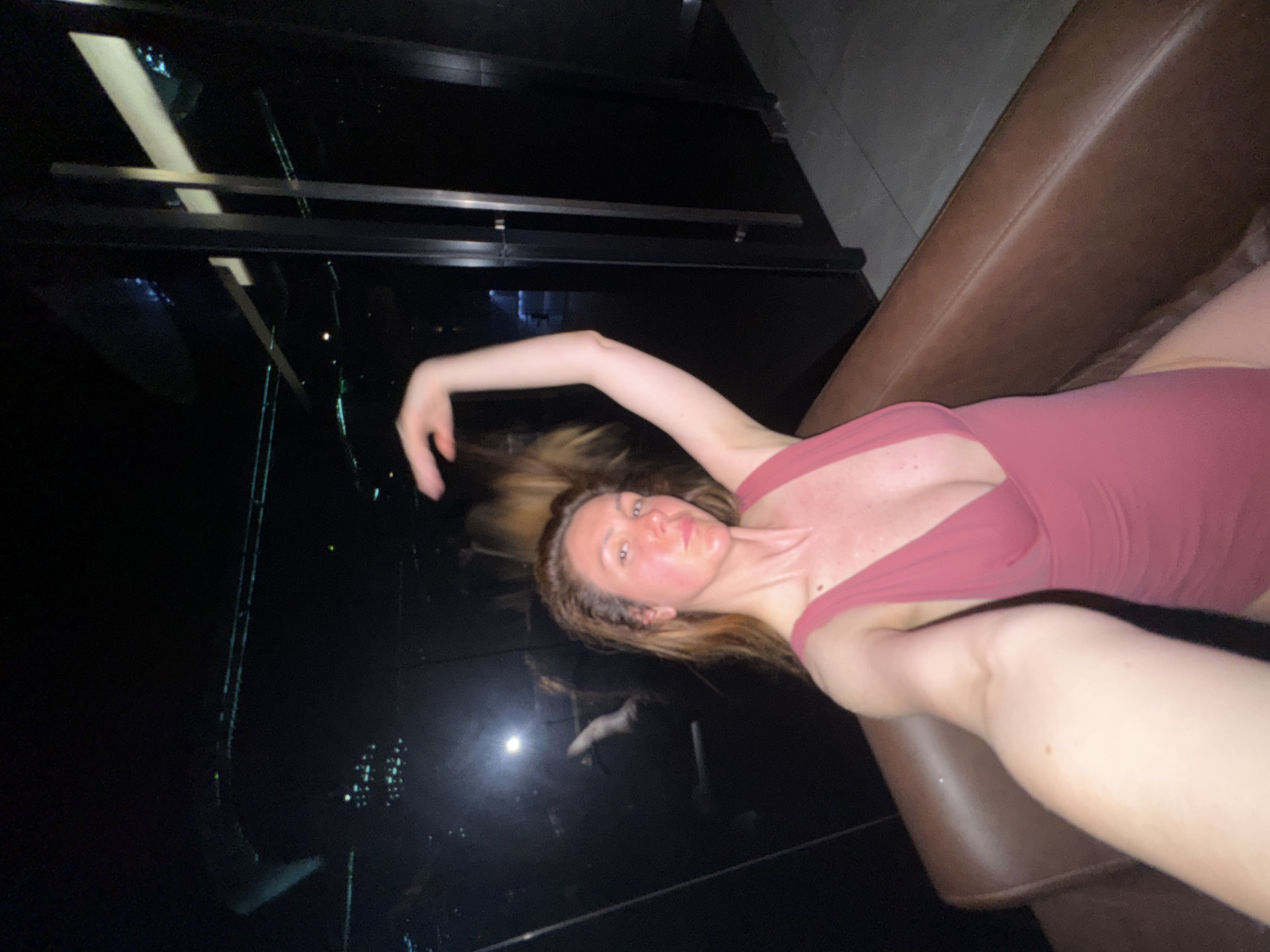 A woman is seated in a brown leather armchair, taking a selfie. She is wearing a deep pink one-piece swimsuit with a plunging neckline. Her hair is a light brown color and is pulled up, with some strands falling around her face and shoulders. Her skin appears flushed, particularly on her cheeks and neck, and there are visible droplets of sweat on her forehead and chest. Her left arm is raised, holding the phone.
The background is dark and appears to be an interior space with reflective surfaces, possibly mirrors or glass. In the reflection, some greenish lights and structural elements can be discerned, suggesting a modern, potentially dimly lit environment. Through a glass partition to the right, a dark corridor or room is visible, with a sliver of a grey tiled floor in the foreground. The overall impression is that of a nightclub, lounge, or a similar establishment in the city of Sofia, Bulgaria, at night.