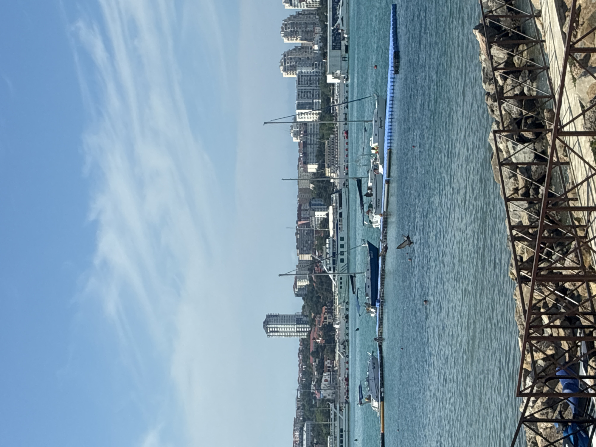 The image depicts a clear day with a blue sky and scattered white clouds over the city of Gelendzhik, Russia. The scene overlooks the Black Sea, with a prominent cityscape in the background. The city features a mix of modern, multi-story buildings and older, low-rise structures, many of which are situated on a hill. A tall, modern high-rise building stands out among them.
In the foreground, a wooden railing with a criss-cross pattern separates the viewer from a rocky shoreline. Beyond the railing, a body of calm, turquoise water is visible. Several boats are moored in the water, including sailboats and motorboats, some docked at a blue floating pontoon. One sailboat has the name "AquaRina" visible on its hull. Two people are visible in the water, and a bird is in flight above the surface. The overall impression is a peaceful day at a coastal resort.