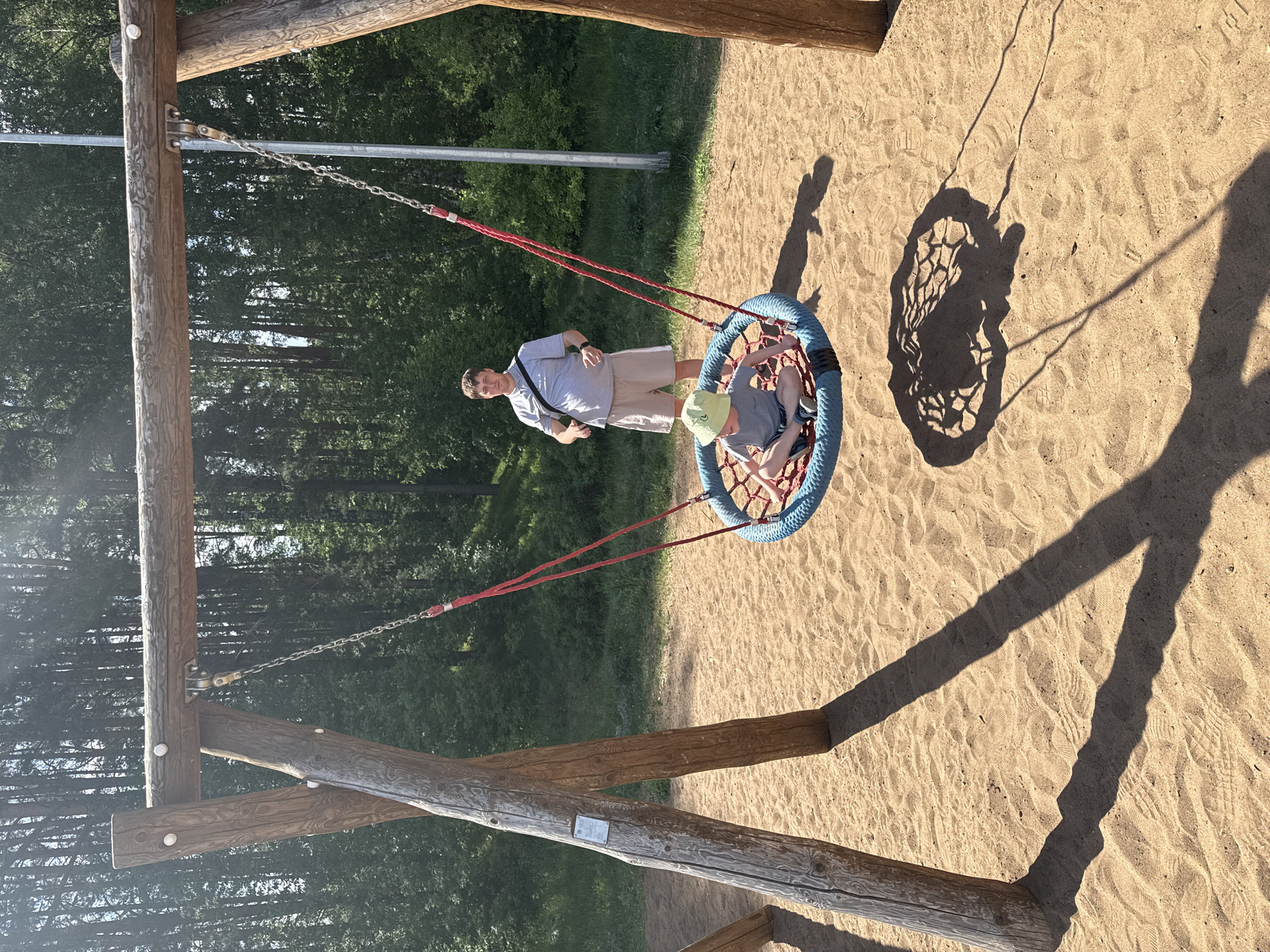 A man stands next to a basket swing holding a child in it. The swing is located on a sandy playground in the city of Chaikovsky, Russia, at latitude 56.77611 and longitude 54.14694. The man is wearing a light-colored short-sleeved shirt, khaki shorts, and a watch on his left wrist. A lanyard hangs around his neck. The child is wearing a grey t-shirt, dark shorts, a green hat and shoes. The swing has a blue rim and a net-like seat with red rope connecting it to the metal chains. The swing set is made of wooden logs. The background includes trees and vegetation. Long shadows are cast on the sand.