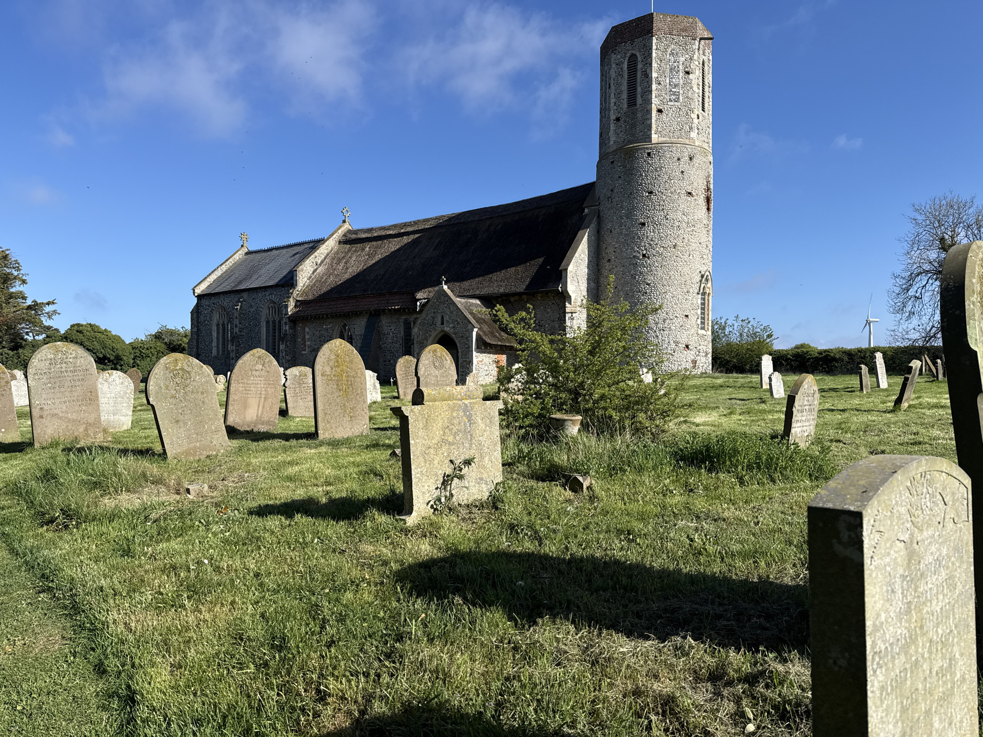 The image displays a historic church and its surrounding graveyard under a bright blue sky with sparse white clouds. The church, presumably in Great Yarmouth, United Kingdom, features a prominent, tall, round stone tower on its right side, characteristic of some British churches. The main body of the church has stone walls and a roof that appears partially thatched and partly covered in darker slate, with decorative crosses on the gables.
The foreground and mid-ground are dominated by a green, grassy churchyard dotted with numerous weathered gravestones of various shapes and sizes. Long shadows are cast across the grass by the gravestones and potentially the church, indicating bright sunlight and possibly morning or late afternoon.
Visible text on a gravestone in the front-left includes partial inscriptions such as "...ES OF HENRY," "MARY CHUBBOCK," and the year "1880." Another stone further back appears to have "MARY ANN" and "JAMES CHUBBOCK 1890" inscribed.
In the distant background, to the far right of the church and beyond a line of trees, a modern white wind turbine is visible against the sky, providing a contrast between the ancient structure and contemporary technology. Small dark specks, likely birds, can be seen flying high in the clear sky. The scene conveys a sense of peaceful history.