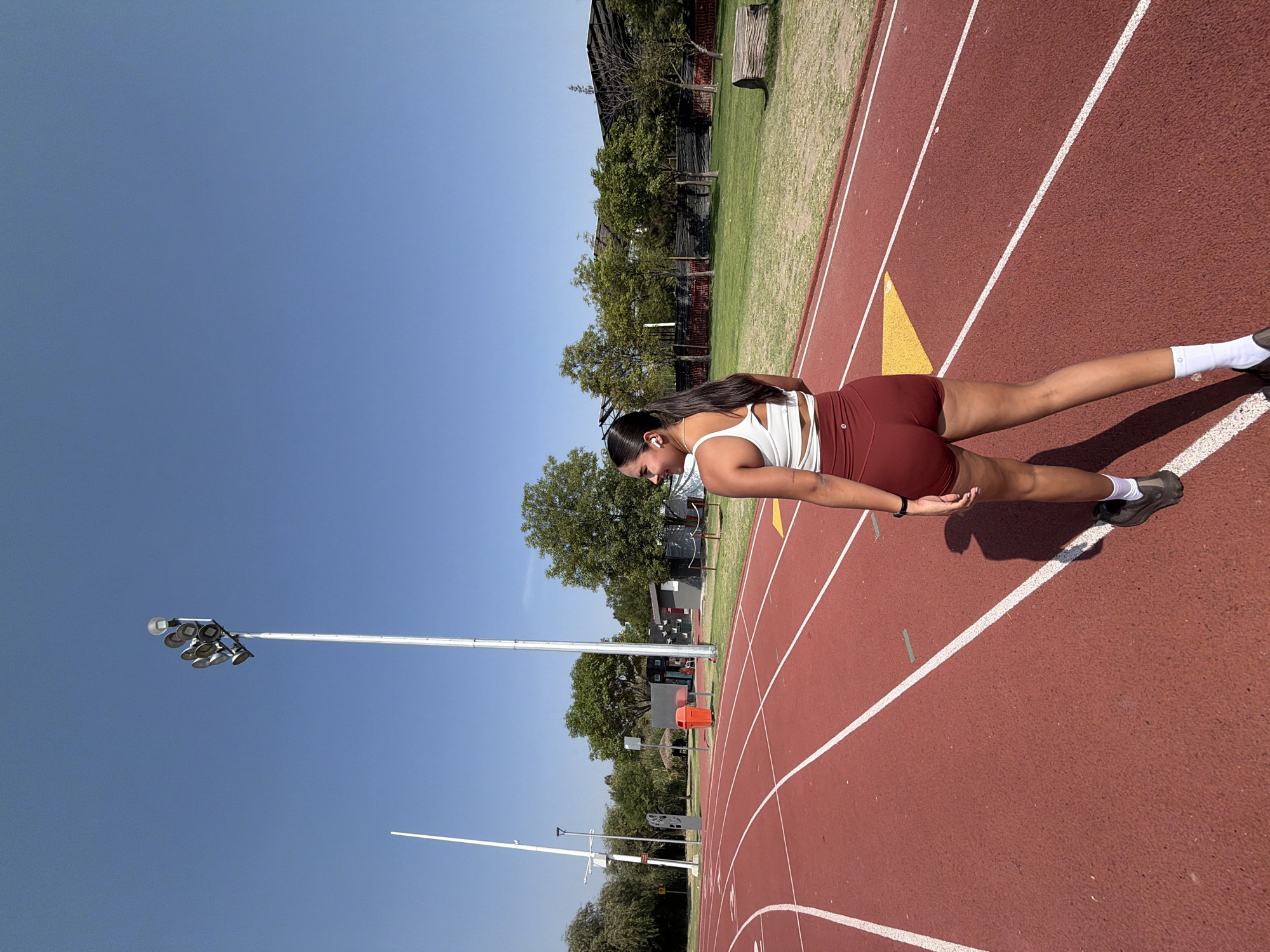 A medium shot depicts a young woman on a track field in Puebla, Mexico. She is clad in a white workout tank top and maroon shorts. The woman is wearing white earbuds and has a dark-colored watch on her left wrist. She has long, dark hair. The running track is red, with white lines delineating the lanes. A yellow triangle is painted on the track. In the background, there is a grassy area, trees, and structures resembling stadium seating. A tall light pole with multiple lights is visible against the clear blue sky.
