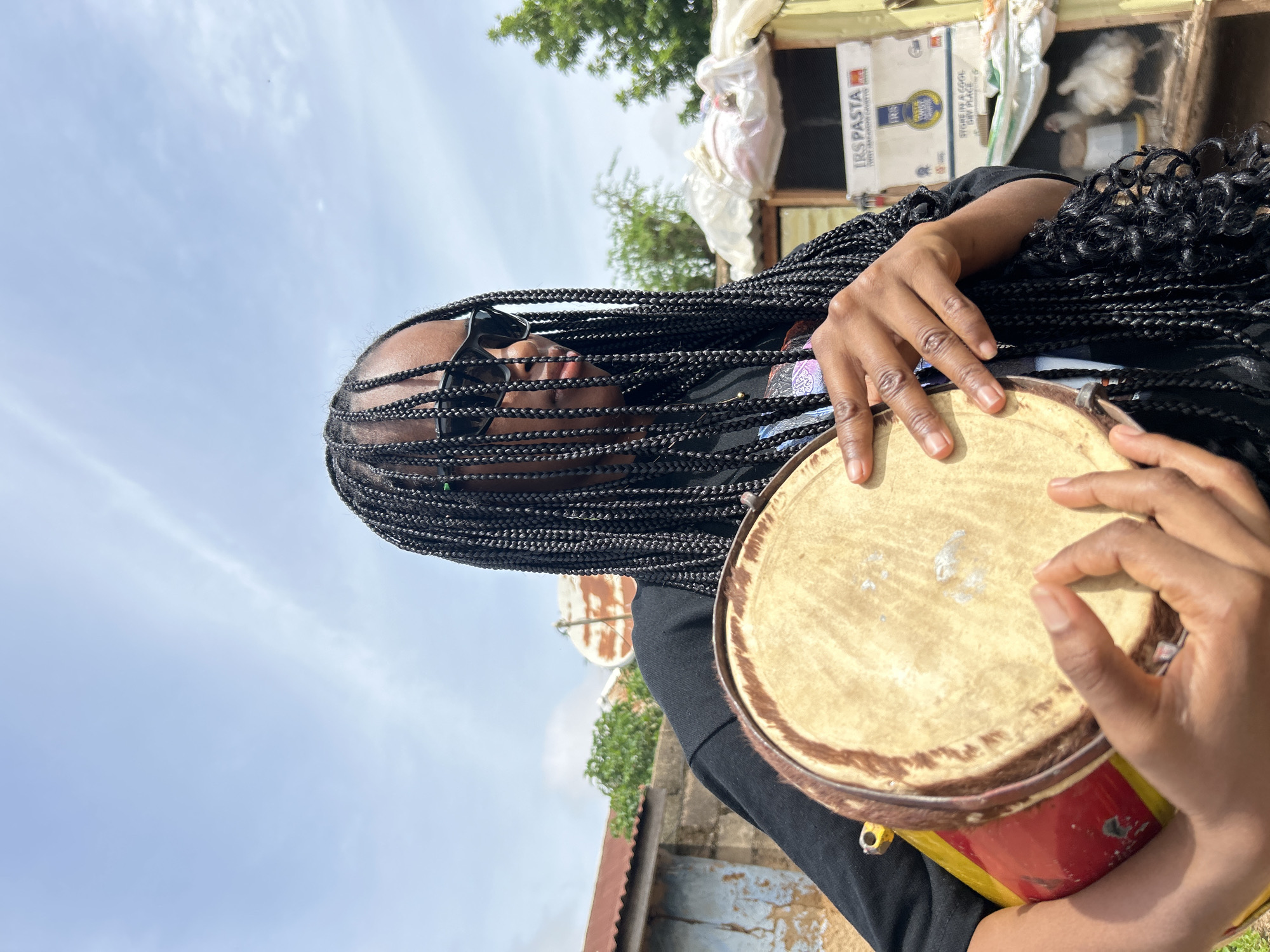The image shows a young Black woman with long, dark braids and black sunglasses holding a small, round drum. She is wearing a black t-shirt. The drum has a light-colored skin stretched over one end and a red and yellow painted body. Her hands are positioned on the drum, suggesting she is about to play it or has just finished playing.
The background is slightly out of focus but shows an outdoor scene that appears to be in a somewhat rural or informal urban setting. There are corrugated metal roofs visible, a rough brick wall, some greenery, and a structure that looks like a market stall or an outdoor storage area. In the background of the stall, there are boxes labeled "IRS PASTA" and what appear to be chickens in a coop. The sky is blue with some wispy clouds.
Given the "IRS PASTA" branding, this scene is likely located in Nigeria, and the mention of Jos, Nigeria in the prompt indicates that the photograph was taken in Jos, a city known for its cooler climate and agricultural produce, though also subject to various economic and social conditions reflected in the informal nature of the background.