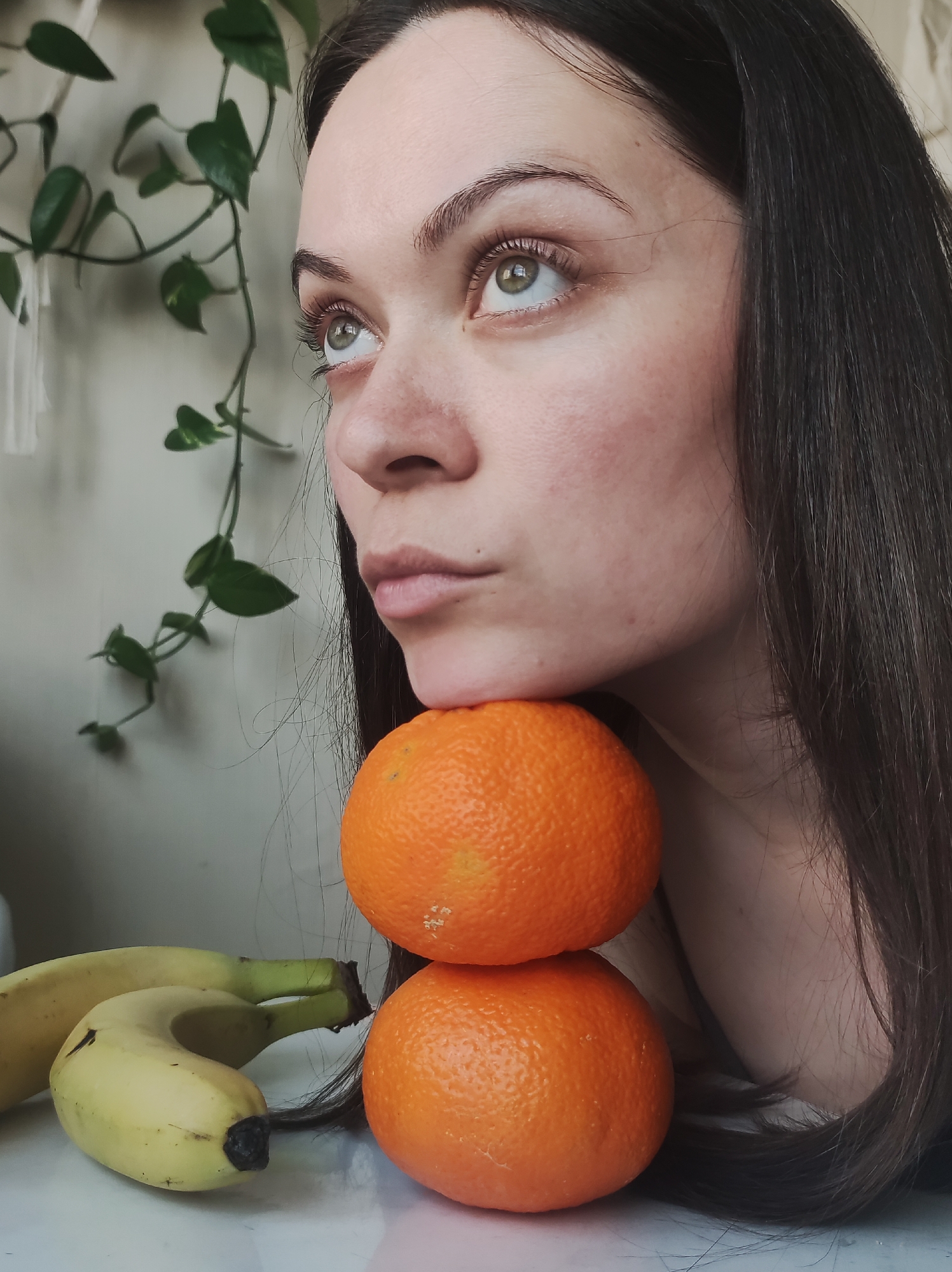 A close-up, eye-level indoor shot shows a woman posing with fruit in what appears to be Chaikovsky, Russia. The woman's face, primarily her left profile (viewer's right), and part of her right shoulder and dark, long hair are visible. She has light skin, and her gaze is directed upwards and to the left, conveying a contemplative or reflective expression. Her chin rests on the top of two stacked, bright orange oranges, which are positioned directly in front of her neck and shoulder. Her hair cascades over her right shoulder and arm, partially obscuring the lower orange.
To the lower left of the frame, two yellow bananas with slight green stems and dark tips are visible, resting on a glossy white surface. In the soft-focused background, a green vine plant with heart-shaped leaves hangs against a light-colored, possibly white or off-white, wall. The lighting suggests a bright indoor setting, likely daytime, with soft illumination on her face. No visible text is present in the image.