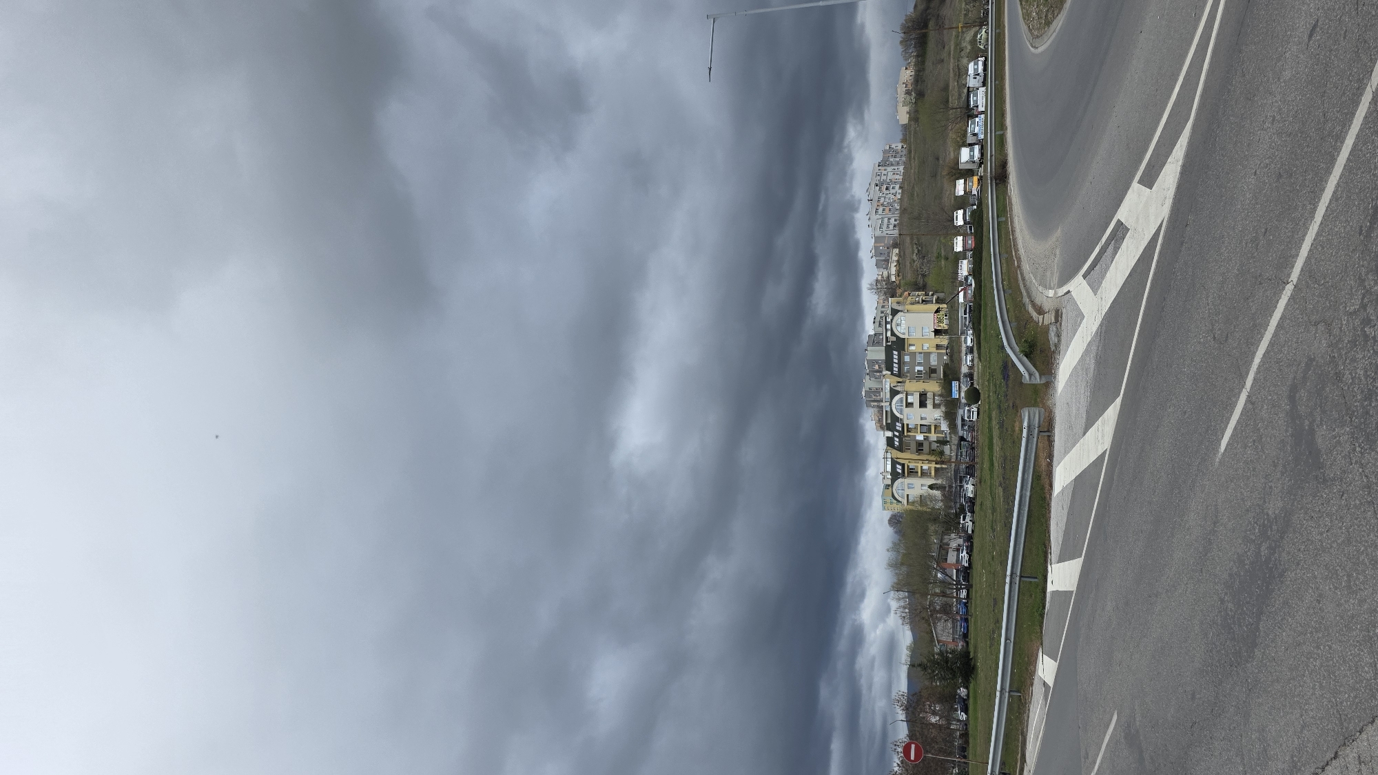 The image shows a street scene in Blagoevgrad, Bulgaria, under a cloudy sky. The foreground is dominated by a paved road, with a median strip featuring diagonal white markings. Alongside the road, there is a guardrail separating it from a grassy area with trees and parked cars. A row of multi-story apartment buildings lines the background, constructed with diverse architectural styles. A streetlamp stands tall behind the buildings. A "no entry" sign is visible at the far edge of the road.