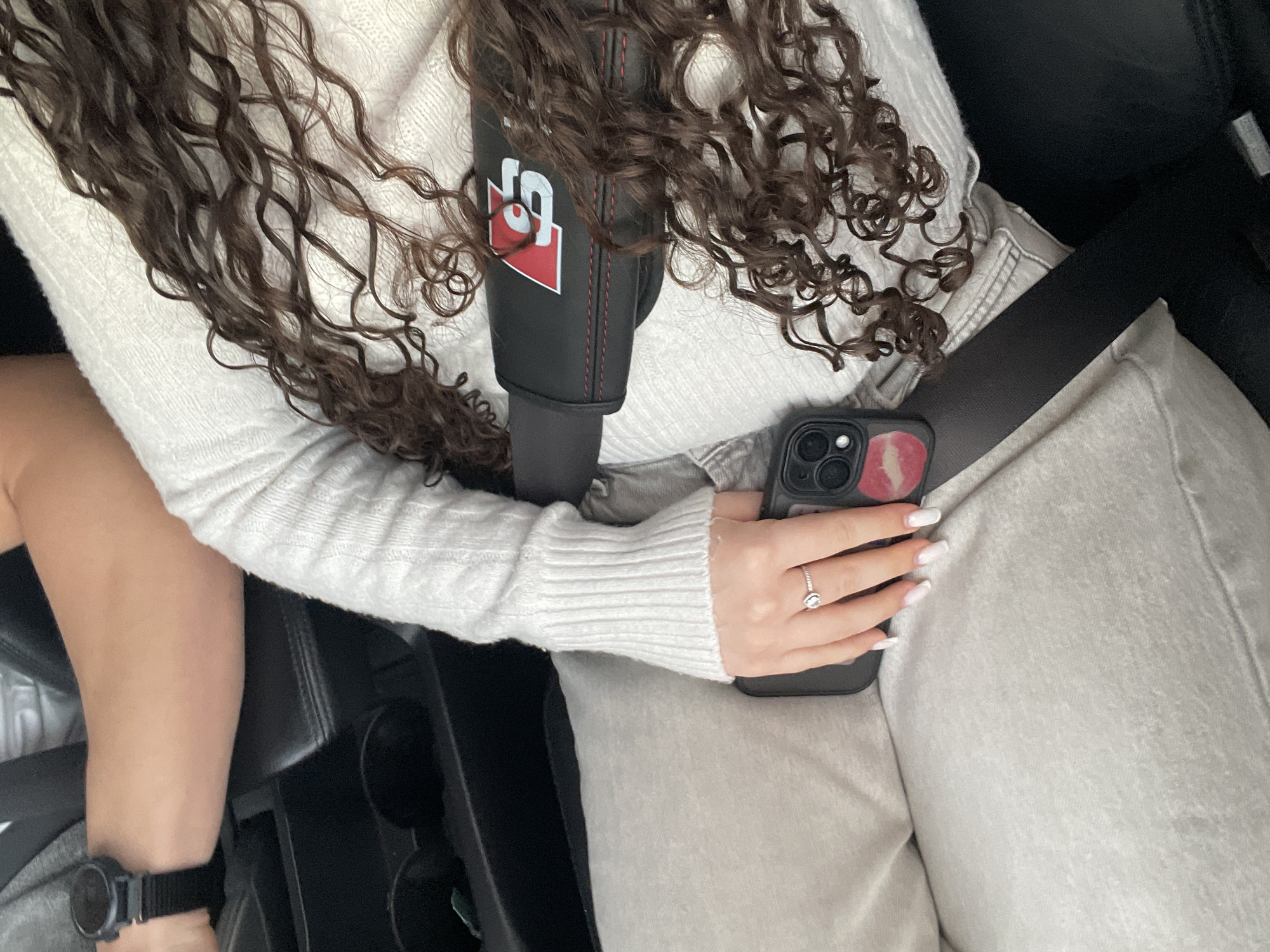 A person sits in a vehicle with a seatbelt fastened across their body. They are wearing a white sweater and light-colored pants. A phone with a red and pink design is held in their hand. The person's hair is long and curly. A black watch is visible on their wrist. The vehicle interior is primarily black. A cup holder and other car features are visible. The city of Zlatograd, Bulgaria, is not discernible from the image.