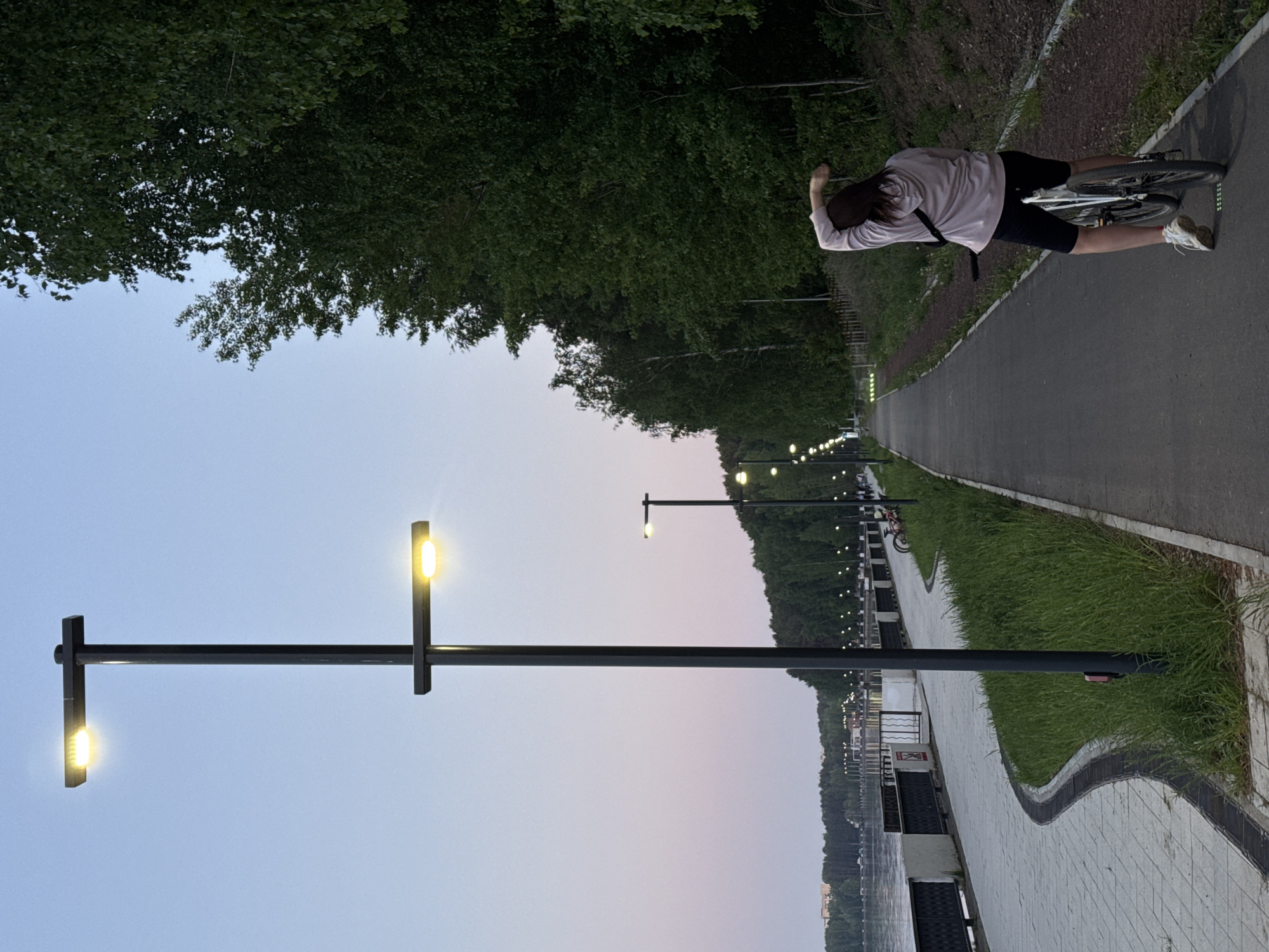 A cyclist in athletic apparel rests on a bicycle on a paved path next to a grassy area, adjacent to a body of water. Streetlights line the path. Trees are visible behind the cyclist and along the path. The location appears to be a waterfront promenade in the city of Chaikovsky, Russia.