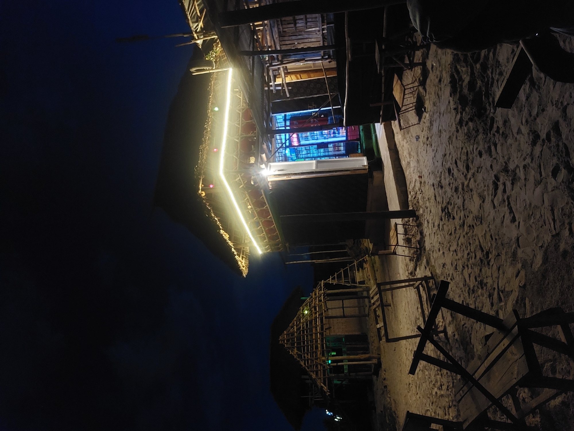 The image shows a night scene in Dar es-Salaam, Tanzania. Two thatched-roof structures are visible, illuminated by string lights along their edges. The foreground shows sandy ground with wooden furniture, including tables and chairs. The sky is a dark blue, suggesting night. The scene appears to be a beachside restaurant or bar. The buildings are wooden frame structures with thatched roofs. One building shows signs of neon lighting.