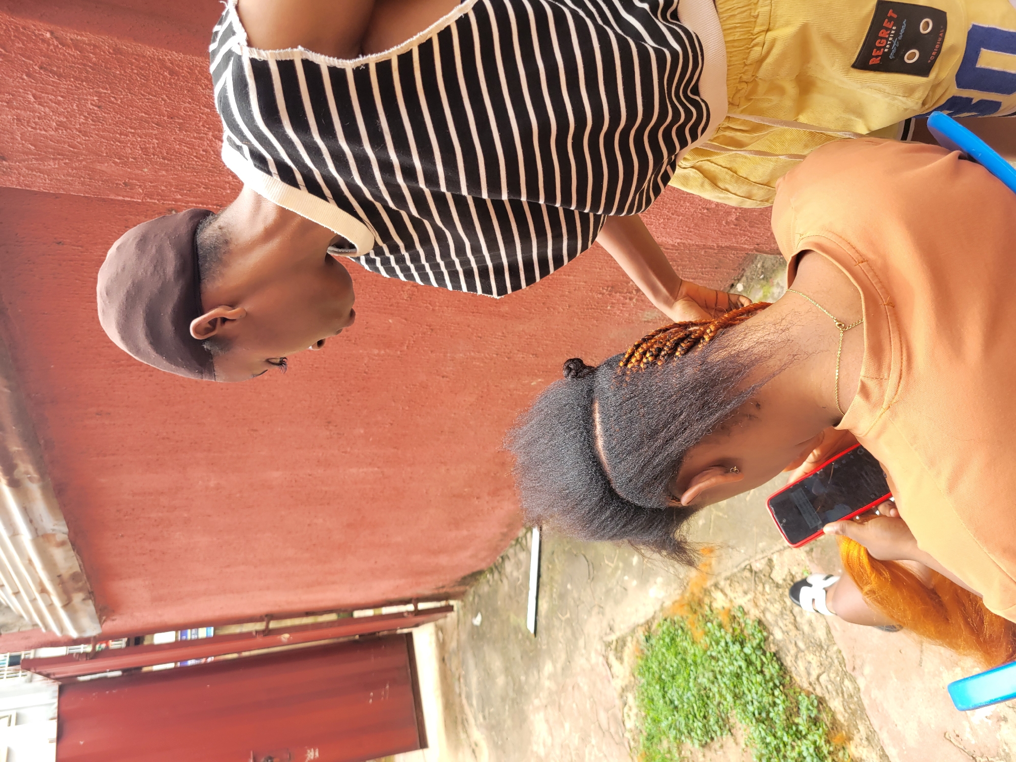 Two individuals are seated outdoors. One person is wearing a black and white striped top and a brown cap. The other person is wearing an orange top and yellow shorts. The person in the orange top is holding a red smartphone. The background shows a red wall and some greenery. The scene appears to be in a residential or outdoor public area. There are some visible text elements on the yellow shorts, including "REGRET NOTHING" and "ORIGINAL". The lighting suggests it is daytime.