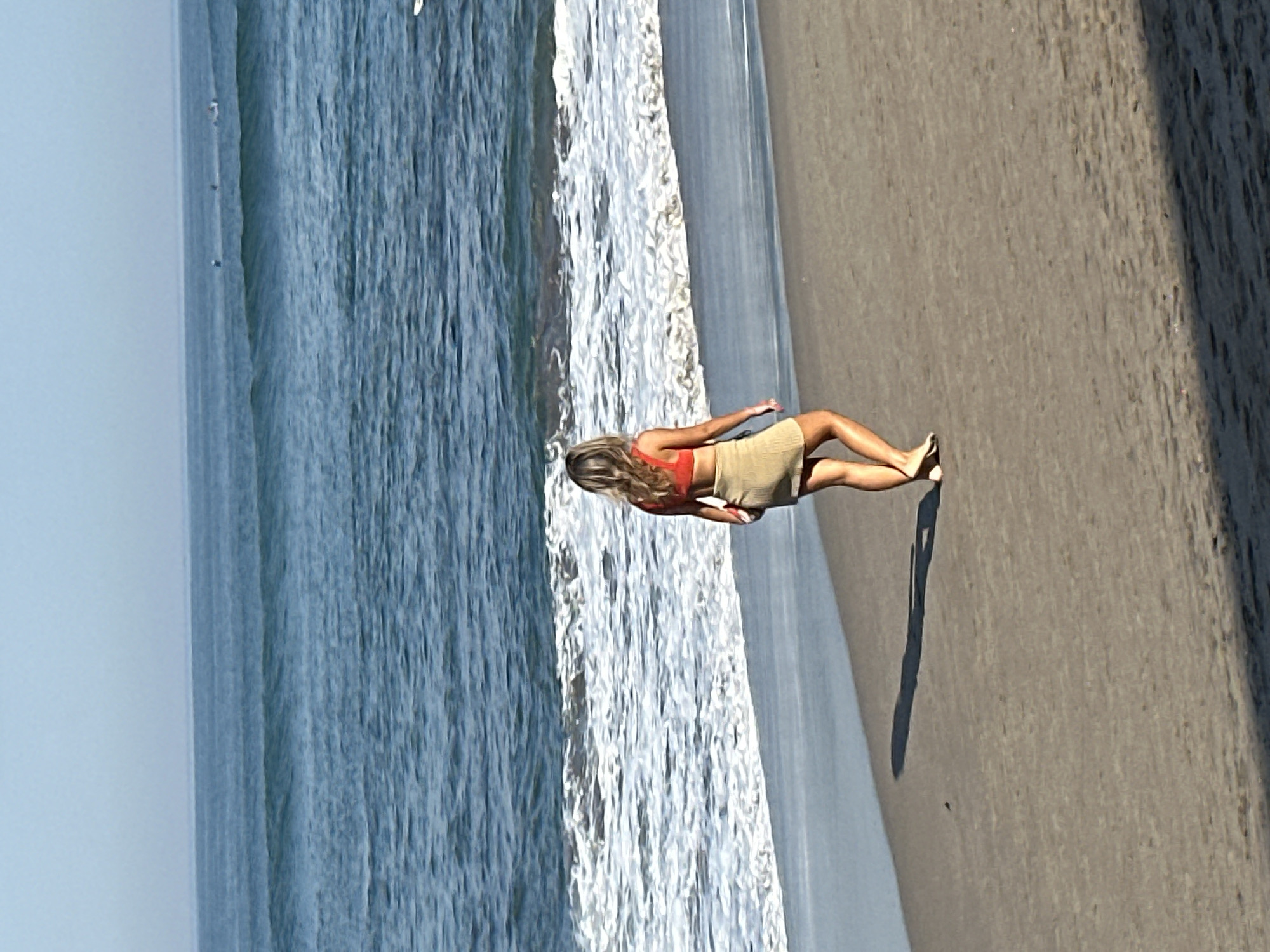 A woman with blonde hair, seen from behind, stands on a sandy beach. She is wearing a coral-colored bikini top and a tan skirt. The ocean with blue waves stretches out before her, meeting the horizon under a hazy sky. There are several surfers in the distance on the water. The woman's shadow is cast long on the sand beside her.