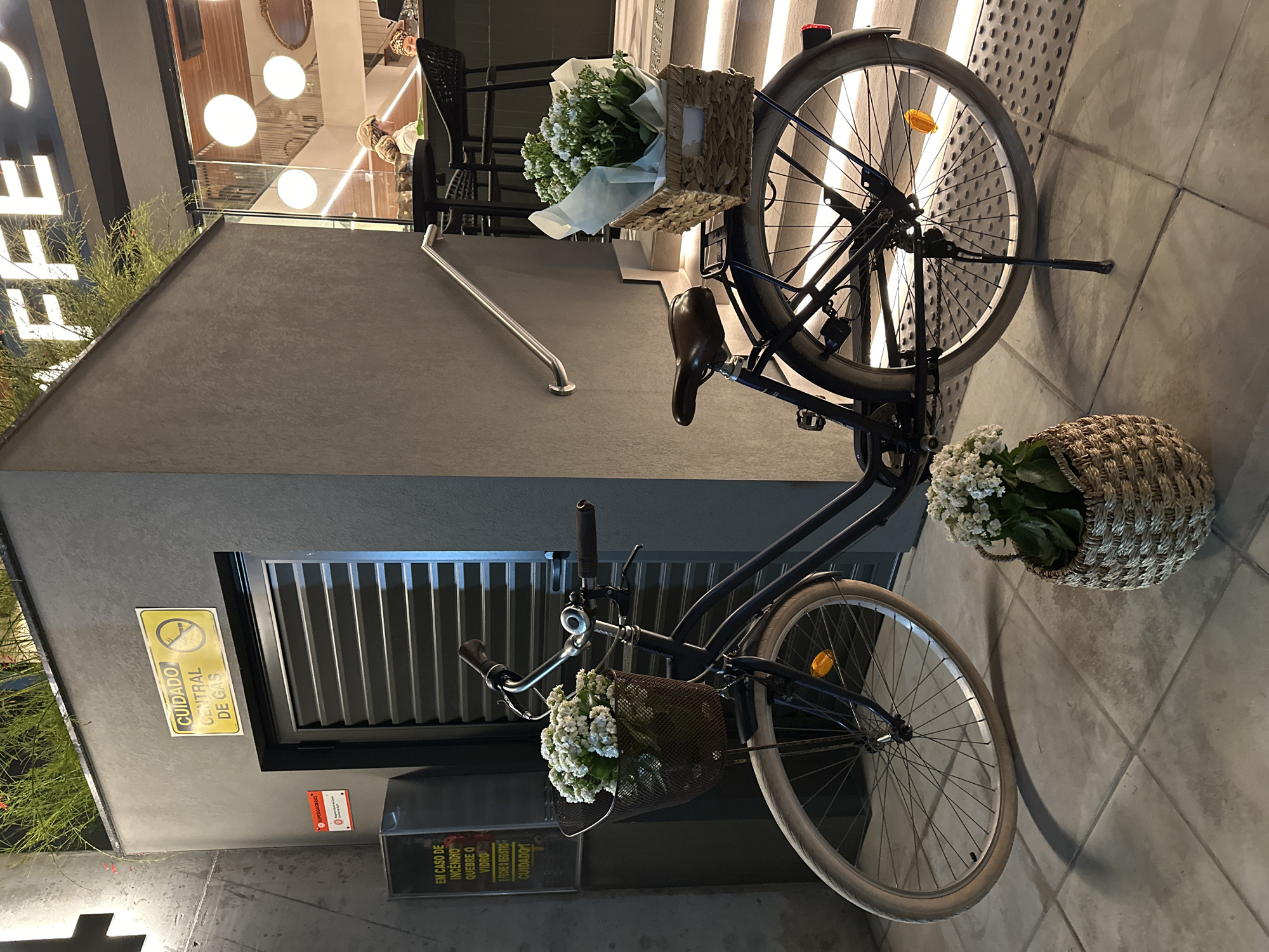 Here's a detailed description of the image:
**General Scene:** The image features a bicycle, decorated with flower baskets, parked near a building in Florianópolis, Brazil.
**Bicycle Details:** The bicycle is a dark-colored model with baskets attached to the front and rear. Both baskets are filled with white flowers. A wicker basket with flowers sits near the back tire on the ground.
**Building Details:** The bicycle is parked next to a gray, modern-looking building. There is a set of grated doors with a sign in Portuguese that reads "Cuidado Central de Gas" (Caution: Central Gas). There is a sign about the event of a fire. Above the bicycle, there are stairs with a metal handrail. A glimpse of a cafe or restaurant interior with lighting and people is visible through an entryway.
**Environment:** The ground is tiled in a gray pattern. The lighting suggests it is either early morning, late evening, or an indoor setting with artificial light.
**Location (Based on available context):** The building appears to be in a commercial area, likely near a cafe or restaurant in Florianópolis. A partial view of the word "FFE" vertically aligned as part of a business name can be seen.