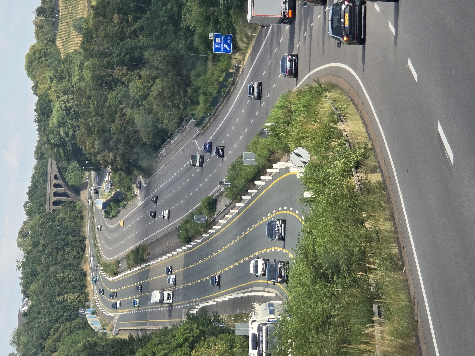 An aerial shot shows a multi-lane highway that curves through a lush, green, forested area. The highway has several lanes separated by yellow lines and white dashed lines. Various vehicles, including cars, trucks, and a white van, are visible on the highway, traveling in different directions. Some of the cars are dark-colored, while others are white or silver. A large white truck with a trailer is also present, as are two smaller white trucks.
On the right side of the highway, there is a blue road sign with white lettering and a parking symbol, indicating a parking area 100 meters ahead. The surrounding environment is dominated by dense trees and vegetation, with a hint of a vineyard on a hillside in the upper right corner. In the distance, a stone bridge with multiple arches spans across the landscape. The sky is a pale blue with some scattered clouds.
The image captures a typical highway scene in a scenic, natural setting, suggesting it is likely located on the outskirts of a city or town, as implied by the presence of a highway and the surrounding natural landscape. The location is identified as Königswinter, Germany.