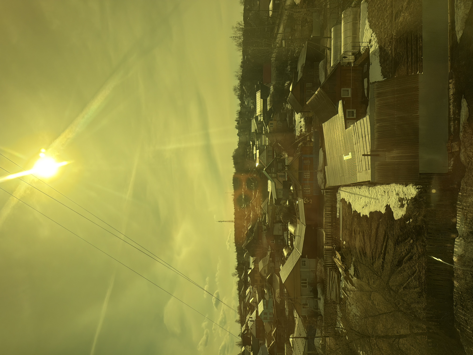 The image presents an elevated view of a residential area, potentially a small town or village, likely located in Chaikovsky, Russia. The structures are primarily single-family homes with pitched roofs, and some appear to be older construction with visible wear and tear. Patches of snow are visible in the foreground and on some rooftops, suggesting a cold climate. The sun is low in the sky, casting long shadows and creating a hazy, golden light across the scene. Overhead power lines are also visible. In the background, there is a treeline marking a wooded area. Silos or cylindrical tanks are present in the middle-ground to the right. A thin tower is visible behind the houses. The overall impression is that of a rural or semi-rural setting in a temperate climate with early spring or late winter conditions. The scene appears to be observed from an elevated position.