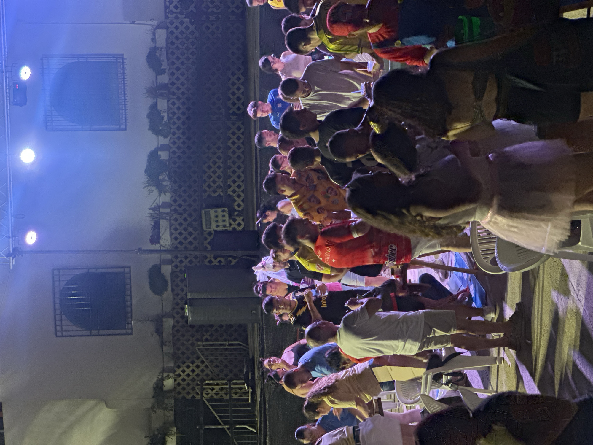 In Blanca, Spain, a crowd of people is gathered outdoors at night, illuminated by blue and white stage lighting. The scene appears to be a social event, possibly a party or concert, as evidenced by the presence of a stage setup with lighting equipment. Many individuals are seen standing and interacting, with some holding drinks and others looking towards a central area, suggesting a focal point of activity. The environment features a building with distinctive arched windows with barred fronts in the background, and a textured lattice-like fence or railing. The ground is paved, and several chairs are visible, indicating a seating area. The attire of the attendees is varied, ranging from casual wear to what might be considered costumes, such as a person dressed in a red outfit with blue overalls, resembling a character. The overall impression is one of a lively, communal gathering under artificial lights.