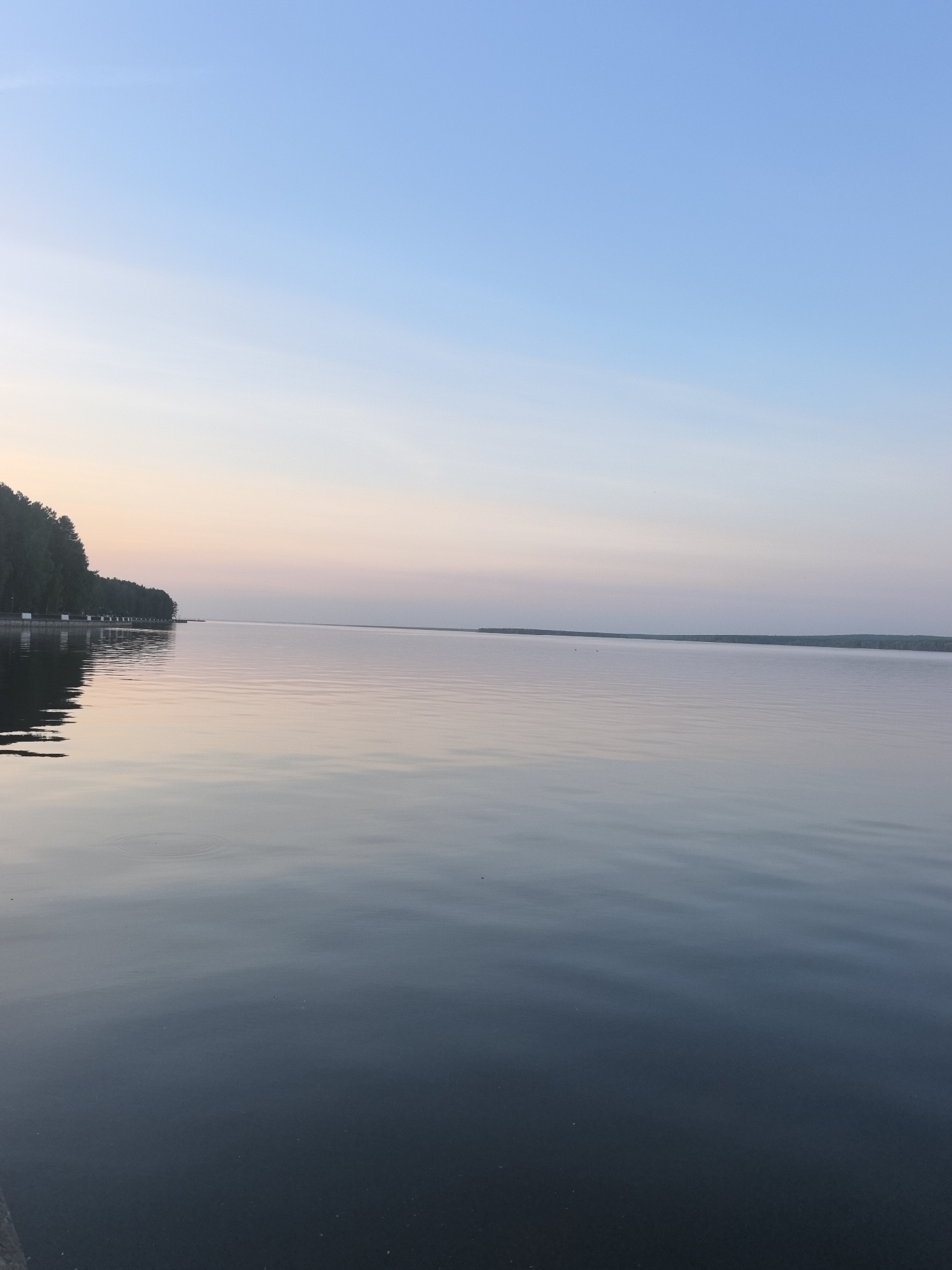 The image depicts a calm body of water, likely a lake or wide river, at dusk or dawn. The sky transitions from a light blue at the top to a soft pink and orange hue near the horizon.
A treeline is visible on the left side of the image, reflected in the water. A distant, low-lying landmass is visible across the water in the middle ground. Small ripples disturb the surface of the water, and the color of the water graduates from light blue-grey where it reflects the sky to a darker blue-grey in the foreground.
Based on the prompt that this image is from the city of Chaikovsky, Russia, the body of water is likely the Votkinsk Reservoir (Воткинское водохранилище).