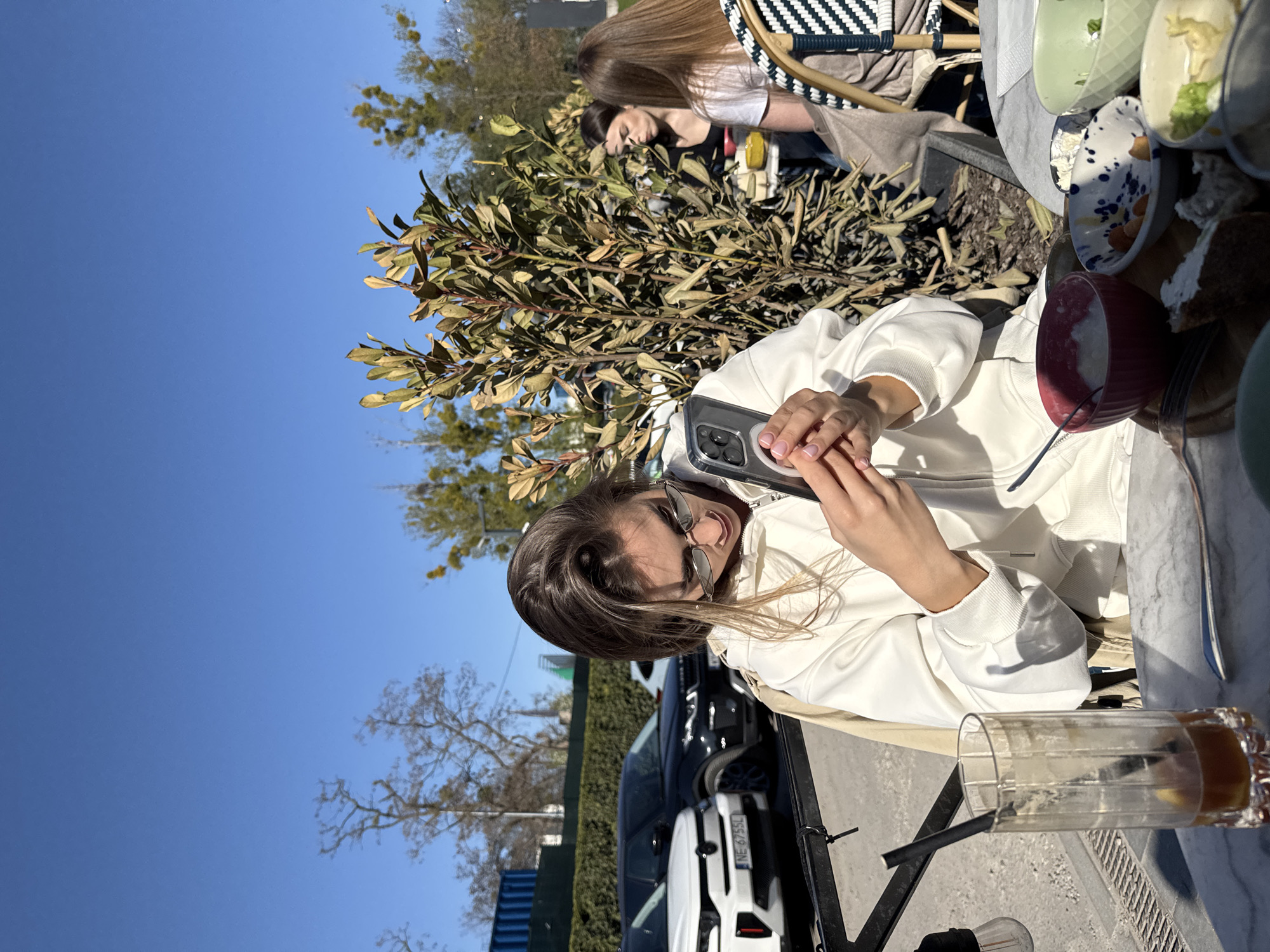 The image depicts an outdoor dining scene on a bright, sunny day. The primary subject is a woman with long brown hair and sunglasses, wearing a white hooded sweatshirt. She is seated at a table, holding a smartphone with a clear case in both hands, actively engaging with its screen, possibly taking a photograph or video. Her light pink manicured nails are visible.
On the table in front of her are several small, colorful bowls, some containing food or food remnants, including a red bowl with a creamy substance, and other bowls in pastel green, cream with blue speckles, and plain cream. A piece of what appears to be cake or bread is also on the table. To her side, a tall glass with a black straw, containing a small amount of liquid residue, suggests a consumed beverage.
In the midground, lush green foliage, likely from bushes or small trees, partially obscures two other individuals seated at a nearby table. Further in the background, an urban environment is visible, featuring parked cars. A white car with the Polish license plate "NE 6755L" is clearly identifiable. The sky above is a clear, bright blue, with some bare tree branches visible against it. The overall setting is a relaxed outdoor cafe or restaurant, characteristic of a casual social gathering in Warsaw, Poland, as indicated by the license plate.
