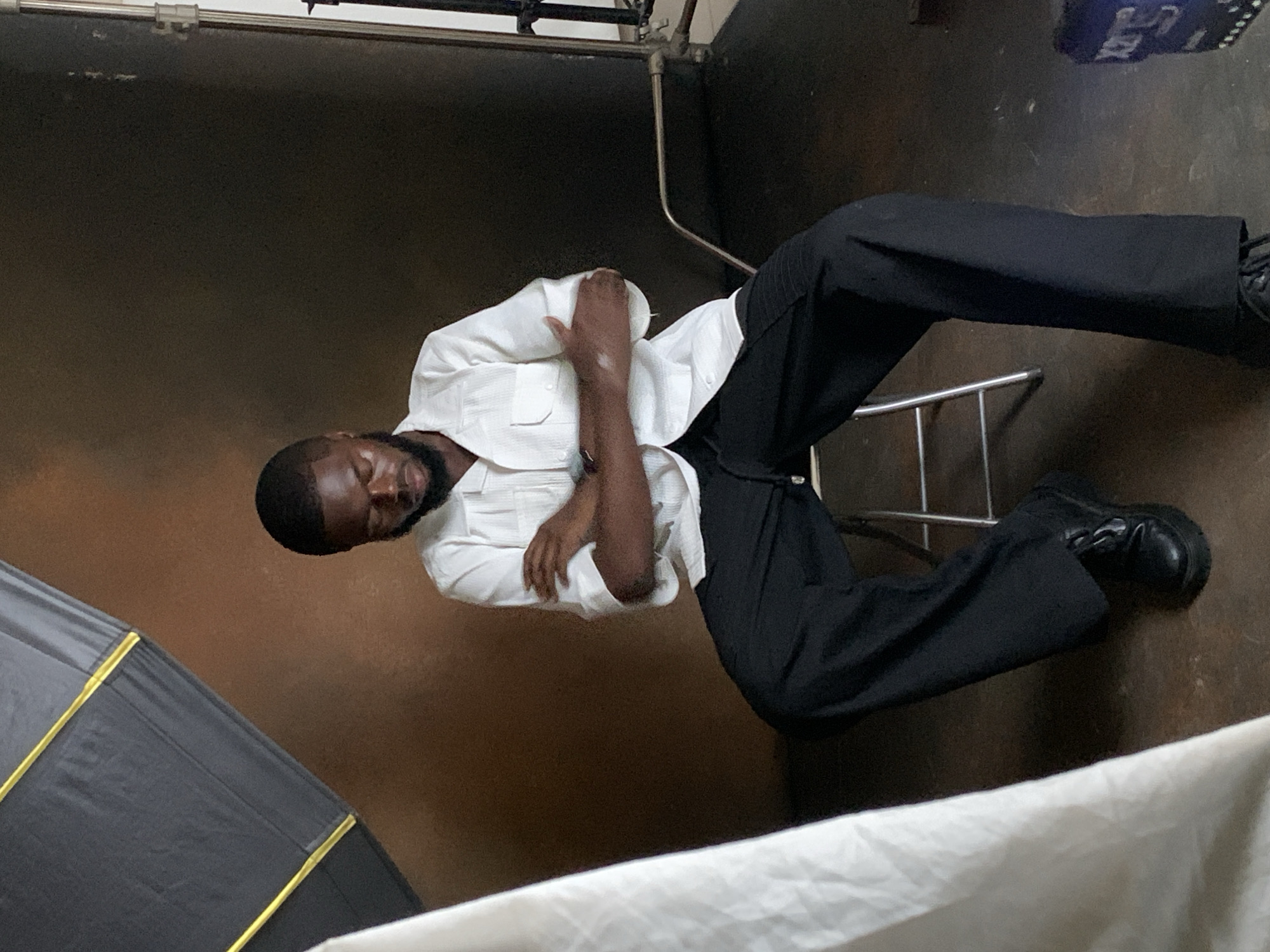 A Black male with short dark hair and a beard sits with his arms crossed on a stool, his head bowed. He is wearing a white short-sleeved collared shirt with two pockets and black wide-leg pants. He has on black shoes with thick soles. The background is a dark brown wall. To the left of the frame, a white fabric hangs, partially obscuring the image. In the upper left corner, a portion of a black and yellow softbox is visible. To the right, a metallic pole is present, and at the bottom right, a blue object with white lettering is partially visible. The scene appears to be a photography studio, potentially located in Jos, Nigeria.
