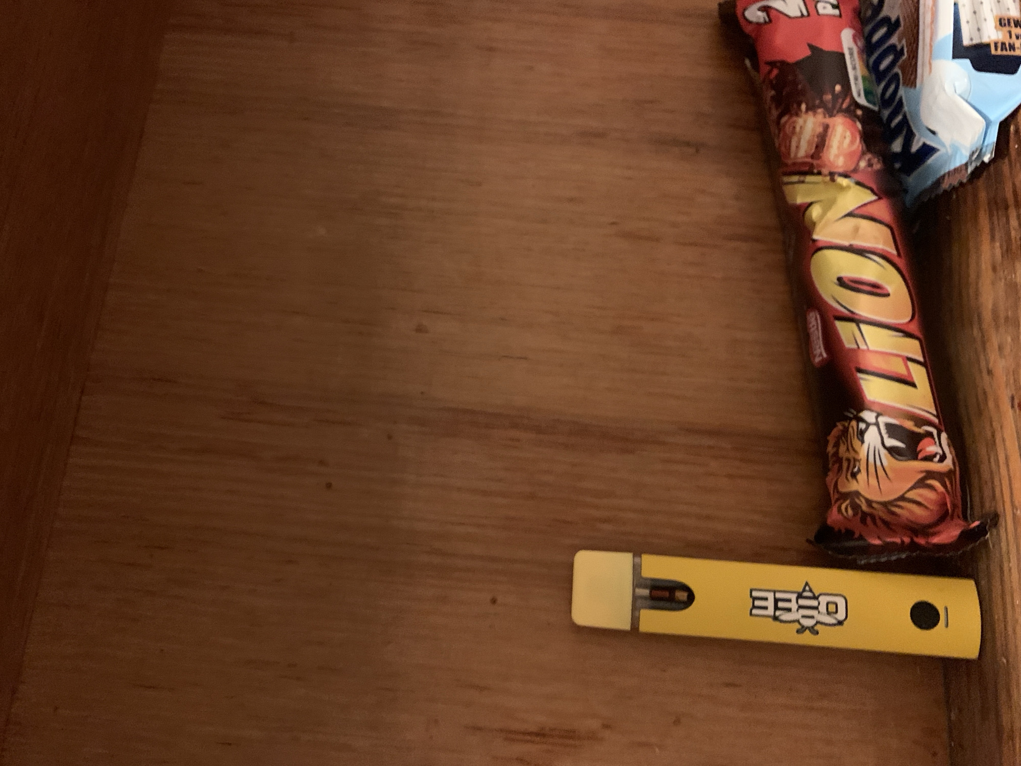The image displays a close-up, top-down view of various items resting on a wooden surface, likely the inside of a drawer or a shallow box, indicated by the raised wooden edge on the right and top. The wood has a visible grain and is a warm brown color.
The main subjects are two wrapped snack bars and a yellow electronic device. On the right side of the frame, there is a red and yellow wrapper for a "LION" chocolate bar, partially obscured by a blue and white wrapper for a "Knoppers" bar. Below these, positioned horizontally towards the bottom right, is a bright yellow, rectangular electronic device. This device has a dark opening on its left side and a small dark circular indicator on its right.
There is no human activity depicted. The scene suggests an indoor setting, possibly a storage area or a personal space where these items have been placed. The lighting appears dim but consistent, without harsh shadows or bright glares.
Visible text includes:
- On the red and yellow wrapper: "LION" in large yellow letters. Part of "Knoppers" is also visible on the blue and white wrapper.
- On the blue and white wrapper: "GEW." and "FAN-" are partially visible.
- On the yellow electronic device: The stylized brand name "QEEG" is prominently displayed in the center.