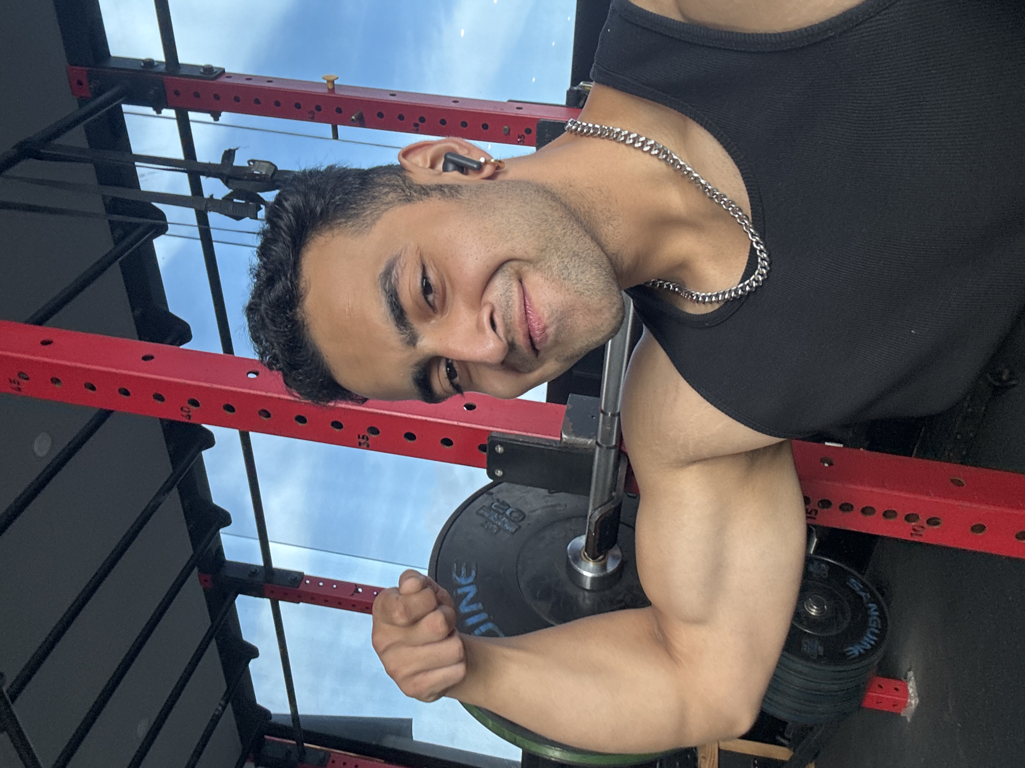 A young man with dark hair and a beard poses in a gym, flexing his bicep. He is wearing a black tank top and a silver chain necklace. He has a black earbud in his right ear. Behind him, a red weight rack is visible, along with a barbell and weight plates. The gym appears to be an indoor facility with large windows showing a blue sky with some clouds. The man's facial expression is a smile. The scene does not contain any identifiable landmarks or elements specific to Kolkata, India.