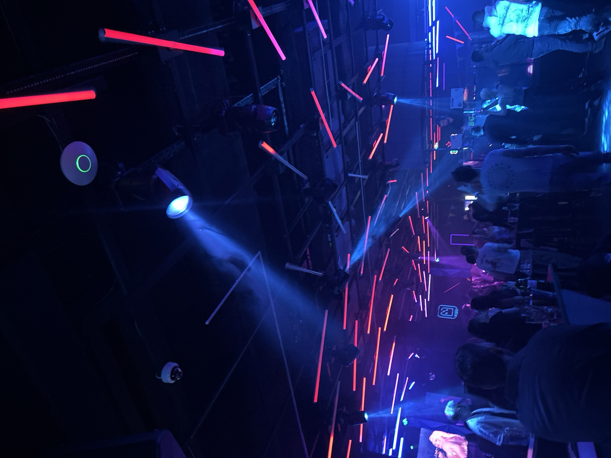 A low-angle, wide shot captures an indoor event, possibly a nightclub or concert, in Sofia, Bulgaria. The dominant colors are blue and pink/red from the extensive lighting system. A trussing system holds spotlights and linear lights, projecting beams into the smoky atmosphere. The crowd is only partially visible, mostly consisting of silhouettes and blurred figures in the foreground. One spotlight emits a prominent blue beam, cutting through the haze. There is a surveillance camera visible in the top left.