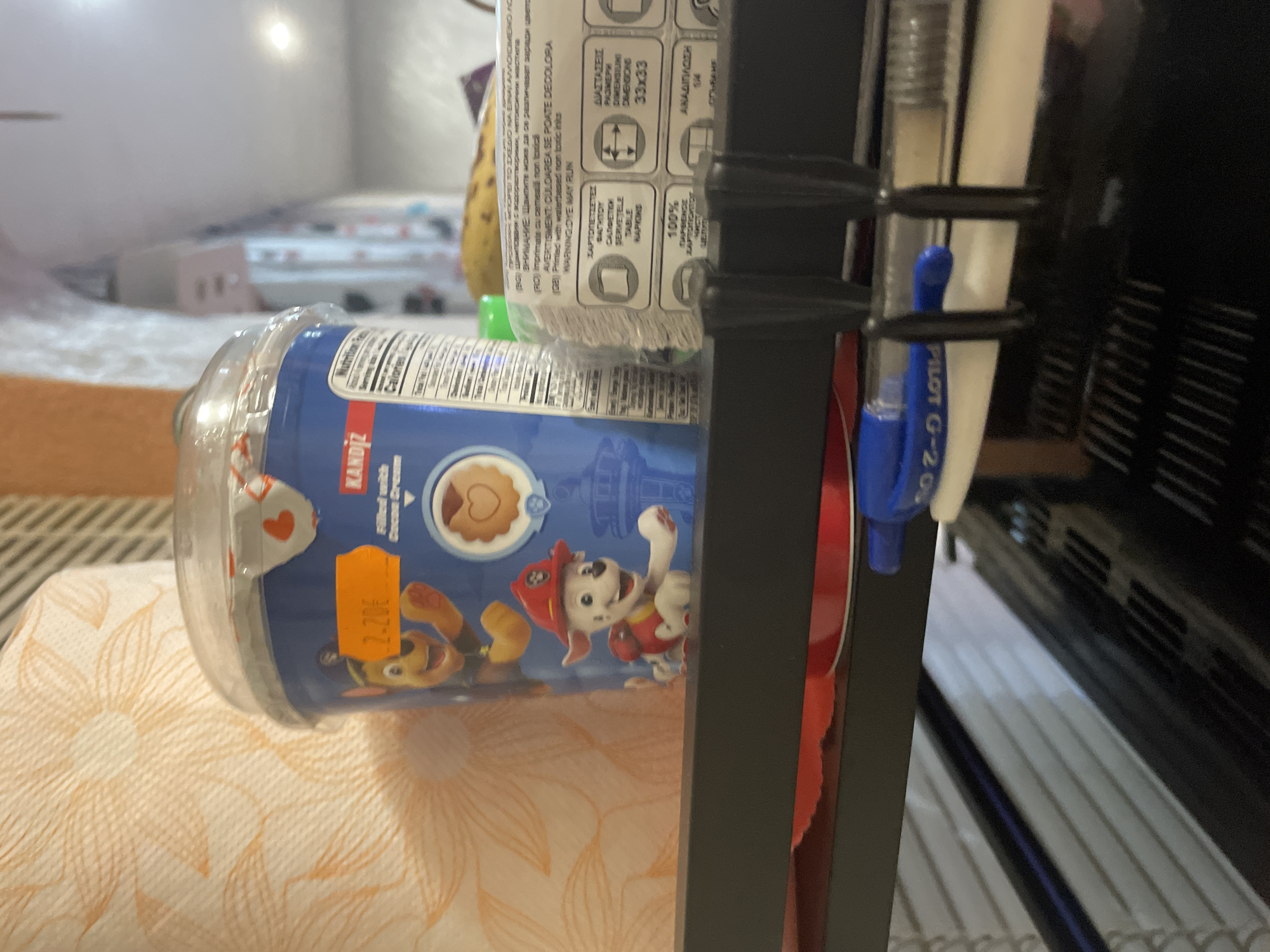 This media file captures a close-up, still-life composition of various items arranged on what appears to be a shelf or surface in an indoor setting in Ihtiman, Bulgaria.
The main subject is a blue cup or container, possibly for cookies or snacks, which is lying on its side. It features illustrations of "Paw Patrol" characters, Marshall and Chase, and has an orange price sticker displaying "2.20€." Visible text on the container includes "KANDIZ," along with partial words like "Filled with Cocoas" and "Calories." A clear plastic lid covers the cup.
Behind the cup, a roll of paper towels or napkins is visible. Its packaging contains multilingual text, including "ΔΙΑΣΤΑΣΕΙΣ," "РАЗМЕРИ," "DIMENSIUNI," and "DIMENSIONS 33x33," indicating its size. Other text includes "100% ΠΑΡΘΕΝΟΣ ΧΑΡΤΟΠΙΟΚΤΟ" (100% Virgin Paper), and warnings like "Printed with waterbased non toxic inks" and "WARNING: DYE MAY RILIN."
To the right, a dark, multi-tiered shelf or rack holds two pens; one is a blue "PILOT G-2 07," and the other is white/clear. A red object is partially obscured behind them. In the soft-focus background, a bed with a patterned sheet is visible, along with a small, light-colored structure, possibly a dollhouse. A yellow banana is also partially seen on the left. The scene is lit by indoor light, and no specific time of day or outdoor weather conditions are discernible.