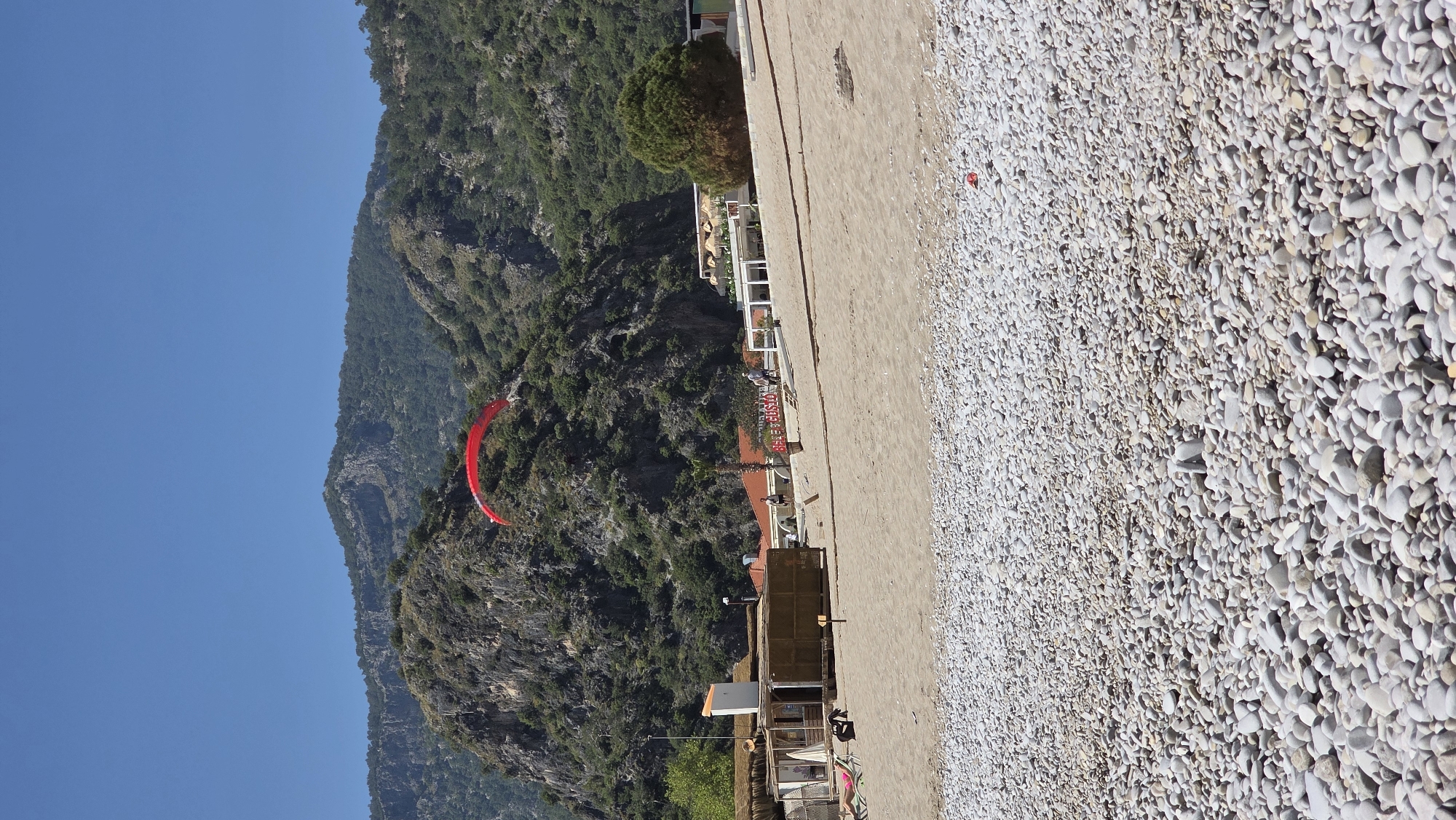 This image, set in Fethiye, Turkey, captures a scenic coastal view on a clear, sunny day. The scene is divided into distinct layers: a pebble beach in the foreground, beachfront buildings in the mid-ground, and majestic mountains under a blue sky in the background.
The foreground features a wide expanse of light-colored, rounded pebbles covering the beach, with a narrow strip of sand visible closer to the buildings. A small red object, possibly a deflated paraglider canopy or a marker, lies on the pebbles near the sandy area.
In the mid-ground, a series of buildings line the beachfront. These structures, possibly restaurants or beach clubs, feature outdoor seating areas with white railings and awnings. One building prominently displays "ALESTA BEACH CLUB RESTAURANT" in red lettering. Several people are scattered around these establishments and on the beach, either seated or standing, enjoying the setting. A large, bushy green tree stands near the buildings, providing shade.
The background is dominated by steep, imposing mountains densely covered with lush green trees and vegetation, rising dramatically against a bright, cloudless blue sky. High above the mountains, a vibrant red paraglider is seen in flight, suggesting popular aerial sports in the area. The overall impression is one of a lively leisure destination, combining natural beauty with recreational activities.