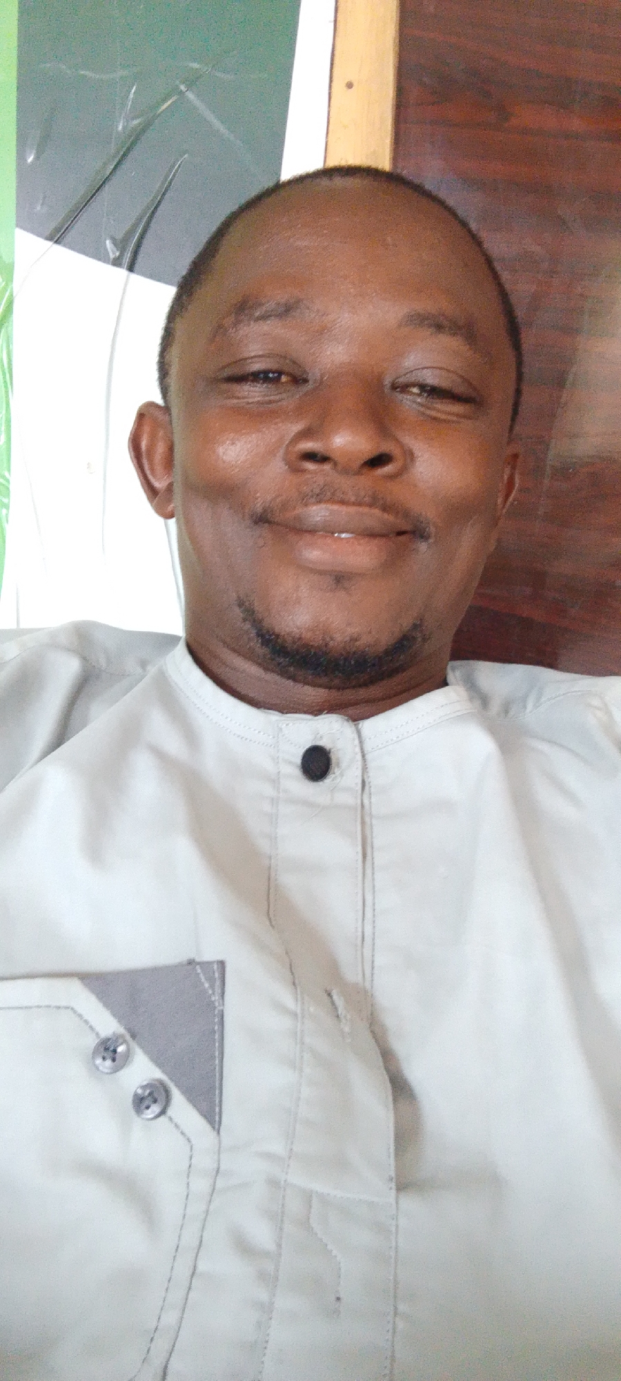 A close-up shot shows an adult male with dark skin and a short beard. He is wearing a light-colored button-down shirt with a gray pocket accent. The man is smiling slightly.
There is no verifiable evidence in the provided image to determine the precise location in the city of Lokoja, Nigeria. The background features a piece of white paper and a wall with light brown wood paneling. Additionally, there is a dark green object to the left of the man's head.