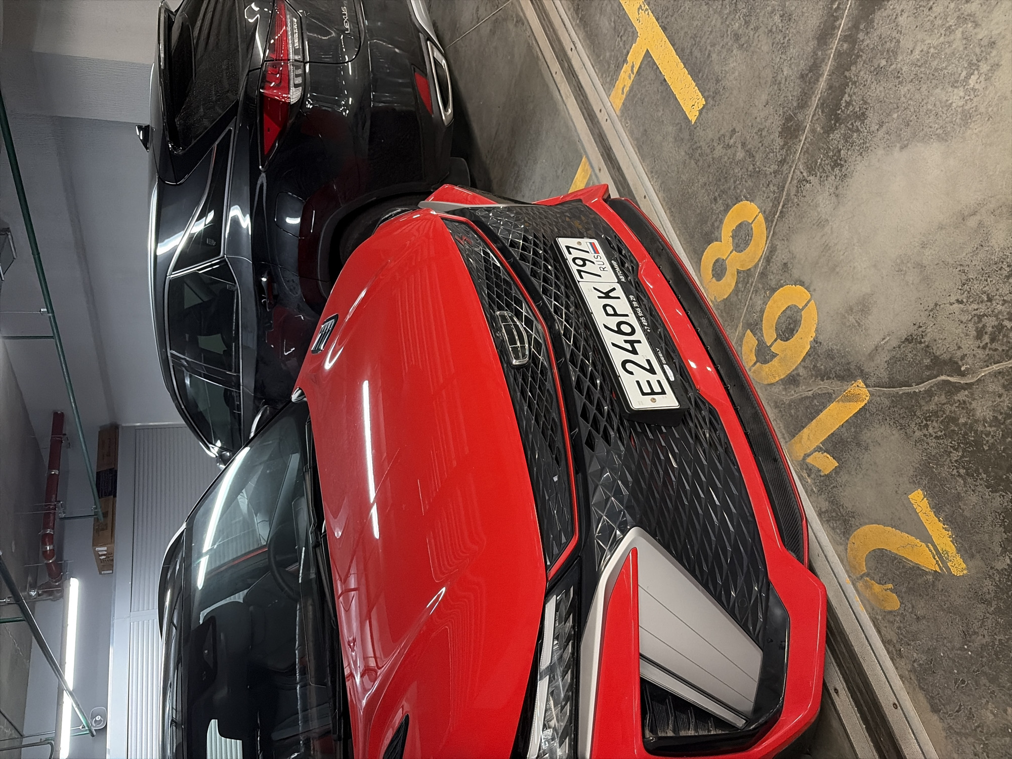 In an underground parking garage in Moscow, Russia, a red Geely car is parked prominently in the foreground, facing slightly left and angled towards the viewer. The car features a distinctive geometric grille and a license plate that reads "E246PK 797 RUS". To the right and behind the red car, a black Lexus SUV is partially visible, its rear end showing a "LEXUS" badge. The garage floor is concrete, marked with yellow painted numbers, including "2" and "68", indicating parking spot numbers. Overhead, fluorescent lights illuminate the scene, and industrial pipes and conduits are visible along the ceiling. The environment appears clean and well-maintained, typical of a modern parking facility. No people are visible in the image.