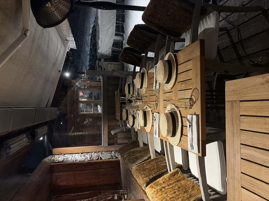 This image captures an empty outdoor dining area, identified as a restaurant terrace in La Clusaz, France, during an evening snowfall. The scene features a long, rectangular wooden table in the center, meticulously set for twelve guests. Each place setting includes a light-colored straw boater hat with a black band, inverted on the table, a white folded napkin with a knife and fork, and a clear drinking glass. Centrally, the table holds a salt and pepper shaker set and a glass bottle.
The chairs along the table are light-toned with white cushions, and many are draped with shaggy brown or amber-colored faux fur throws over their backs. The dining area is covered by an overhead awning structure from which several elongated infrared strip heaters are mounted. A tall, dark, freestanding patio heater with a circular protective grill is positioned on the far right side of the image.
To the left, a rustic wooden wall with stone elements and a prominent horizontal wooden beam defines the interior edge. A transparent barrier separates the covered dining space from the exterior environment. Beyond this barrier, snow is actively falling, evidenced by blurred white streaks against the darker background. The ground outside is covered in snow, and a large white tarp conceals an unidentified object or structure in the middle distance. Distant wooden chalet-style buildings with illuminated windows are visible through the falling snow. No individuals are present in the scene.