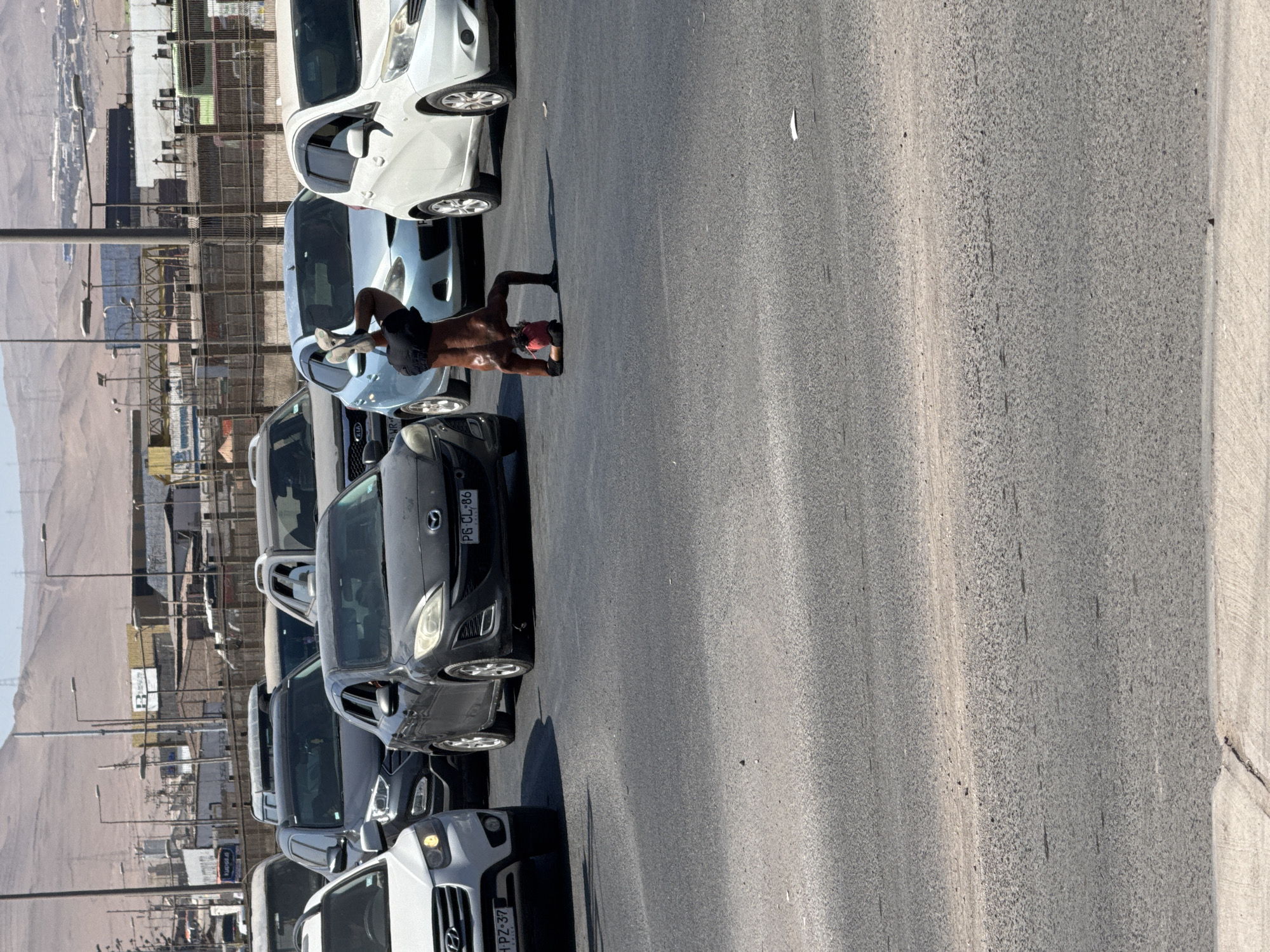 The image captures a daytime urban street scene, location Unknown, Unknown (Chile, based on visible license plates). A dark-skinned individual, shirtless and wearing dark shorts, performs a handstand on the asphalt in the middle of a multi-lane road. His head rests on a small red object, with his hands supporting his inverted body and legs extended upwards, feet wearing light-colored athletic shoes. Vehicles are stopped or moving slowly around the individual. In the foreground, from left to right, are the front of a white Hyundai Santa Fe (license plate H PZ-37 CHILE), a dark gray Mazda 3 sedan (license plate PG CL-86 CHILE), a light blue hatchback, and a white Nissan Note. Additional vehicles are visible further back in multiple lanes. The road surface is dark asphalt with faded white lane markings. The background features arid, light-brown hills under a clear blue sky, with various industrial or commercial buildings, utility poles, and an elevated barrier visible between the road and the hills.