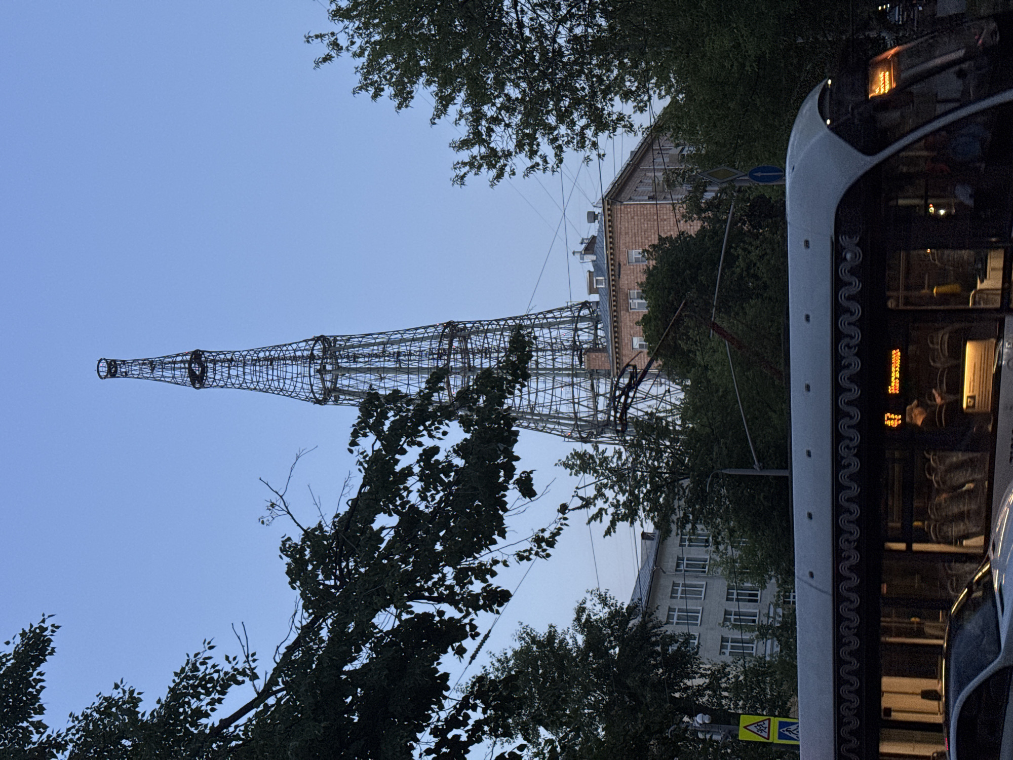 The image shows the Shukhov Tower in Moscow, Russia, which is a hyperboloid radio tower. In the foreground, a tram is visible, with passengers inside. Trees and buildings line the street, and the sky is a clear blue. The tower itself is a tall, lattice structure, with a conical shape. The tram is modern and white, with large windows. The buildings in the background are a mix of architectural styles, and some are residential, while others appear to be offices or public buildings. The overall scene depicts a typical urban landscape in Moscow, with a blend of historical and modern elements.