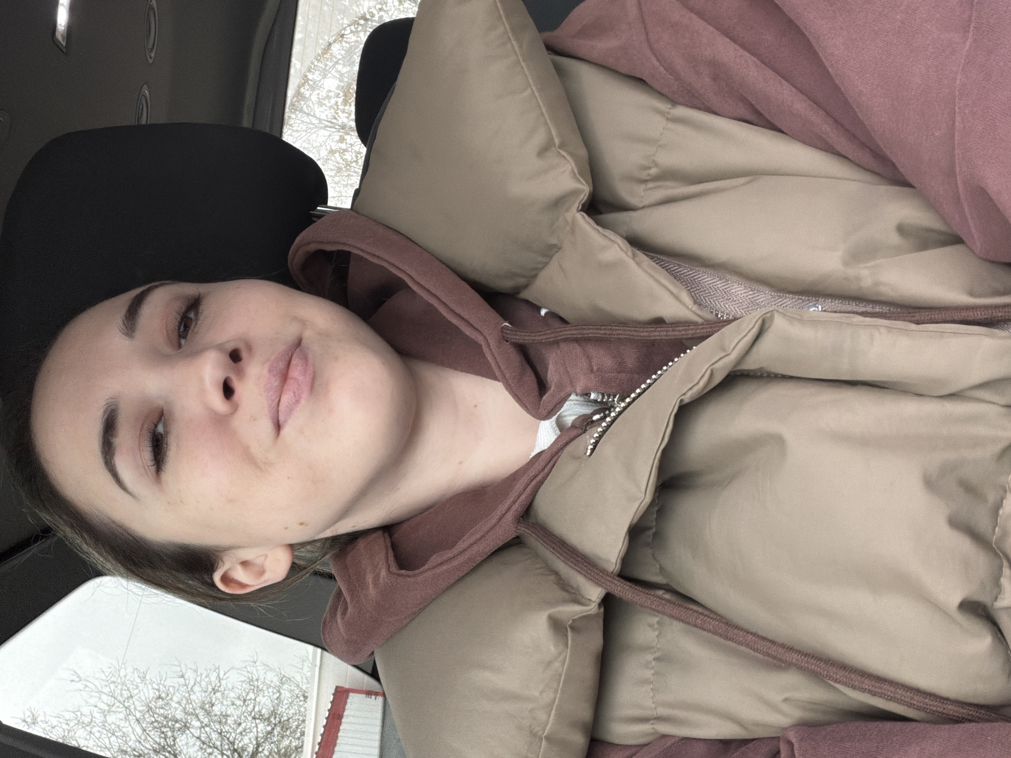 A young woman is seated in what appears to be a car, possibly in the city of Rostov-on-Don, Russia, given the request. She is wearing a brown hoodie and a tan puffer jacket. Part of the car's interior is visible, including the headrest of the seat. Outside the car window, bare trees are visible, with glimpses of buildings, one of which has red and white accents. The lighting suggests it might be daytime. The woman is looking at the camera with a slight smile.