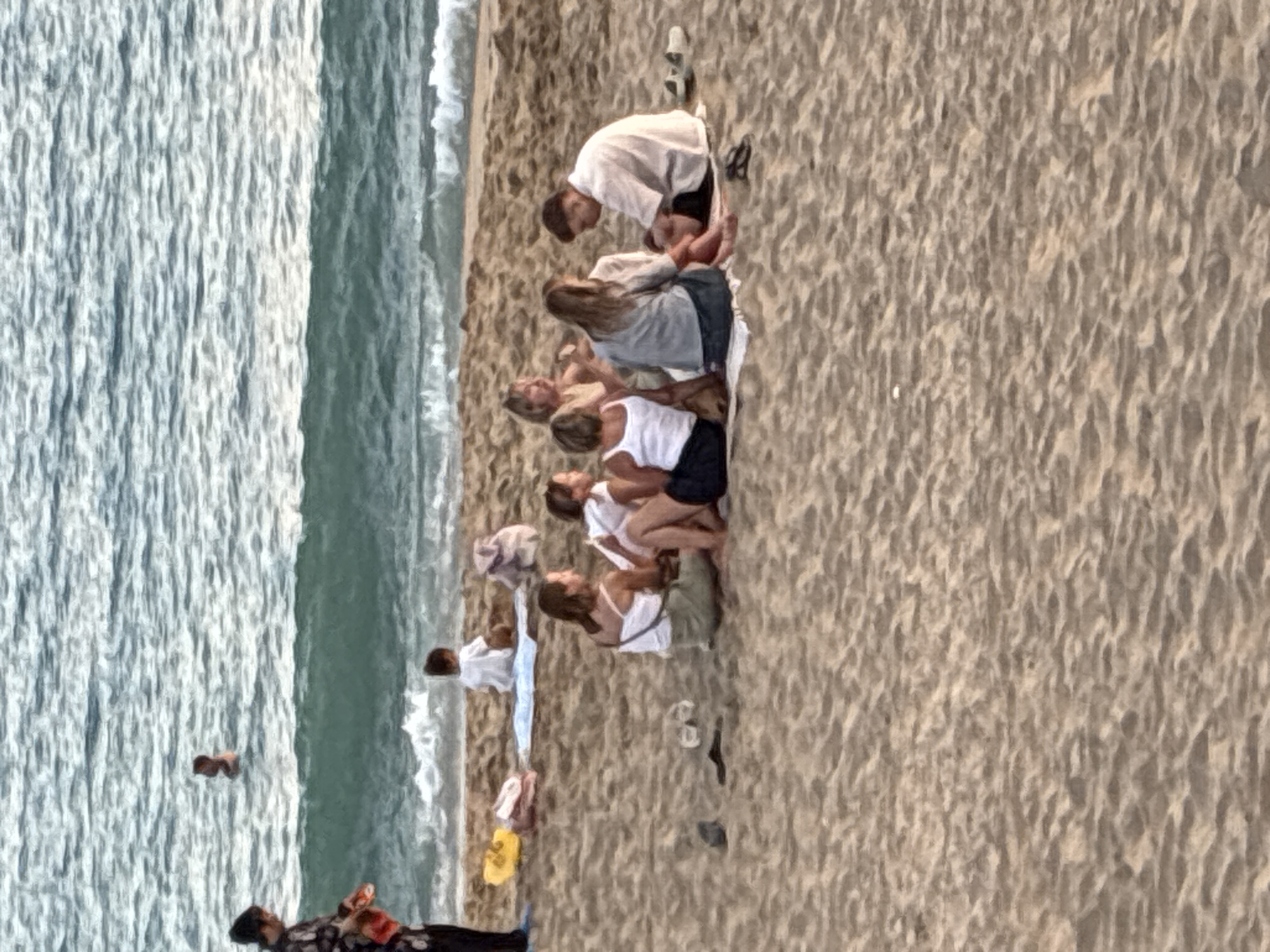 A group of people are gathered on a sandy beach, with the sea in the background. The scene appears to be in Alanya, Turkey, given the context. In the foreground, a group of approximately seven individuals are sitting on towels on the sand. Two are women in white tops and dark bottoms, facing each other. A woman with blonde hair and a beige top is in the center of this group, and a man in a light-colored shirt and dark shorts is seated to her right. Further back, a young child in a white shirt is sitting on a light blue towel, with a yellow object and bags nearby. A person is standing near the water's edge on the left, wearing dark clothing. In the sea, a person is visible swimming in the water. The water is calm with small waves breaking near the shore. The sand is light brown and textured.