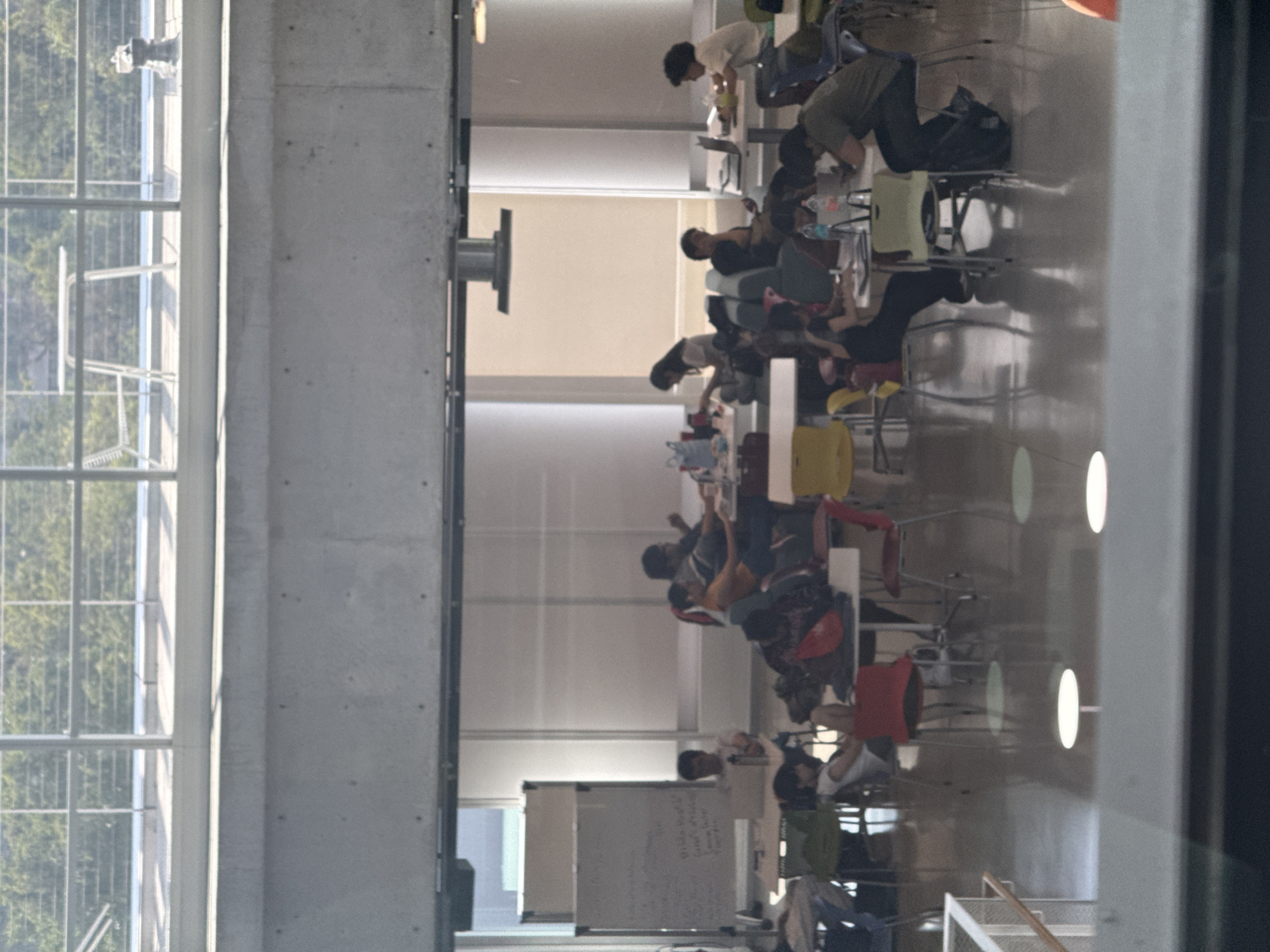 The image depicts an indoor common area, likely a university lounge or co-working space, teeming with people engaged in various activities. The perspective is across the room, which features large, multi-paned windows along the left side, providing ample natural light and a view of lush green trees and distant, blurry structures. A prominent concrete pillar or wall stands on the right.
Numerous individuals, mostly young adults, are seated at different types of tables – some light-colored, some darker – throughout the space. Many are focused on laptops, indicating work or study, while others appear to be conversing or observing. Chairs in vibrant colors like yellow, red, and green, along with some lounge-style seating, are scattered around the tables. Water bottles and other personal items are visible on tabletops.
A notable feature is the highly reflective floor, which mirrors the people, furniture, and several bright, circular overhead lights, creating a dynamic visual effect and adding depth to the scene. In the background, two whiteboards are visible; one contains handwritten text or diagrams, though it is illegible due to the distance and resolution.
The bright natural light streaming through the windows suggests it is daytime. The overall atmosphere appears productive and relatively calm. The modern architecture with concrete elements and large windows, combined with the activities, points to a contemporary educational or collaborative environment.