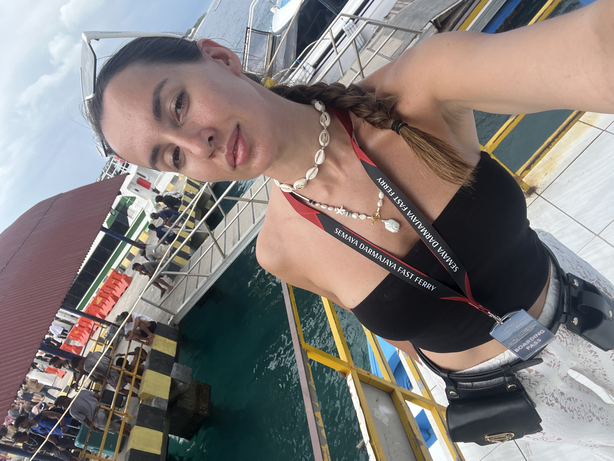 A medium, slightly angled outdoor shot depicts a woman with light skin, brown hair in a braid, wearing a black top, a seashell necklace, and a lanyard with a "Semaya Darmajaya Fast Ferry" label and a "BOARDING PASS." She is wearing white lace pants and a small black purse around her waist. The background shows a ferry dock with yellow and black striped barriers and green water. People are visible on a ferry and a gangway. The location is the city of Unknown, Indonesia. The sky is partly cloudy.