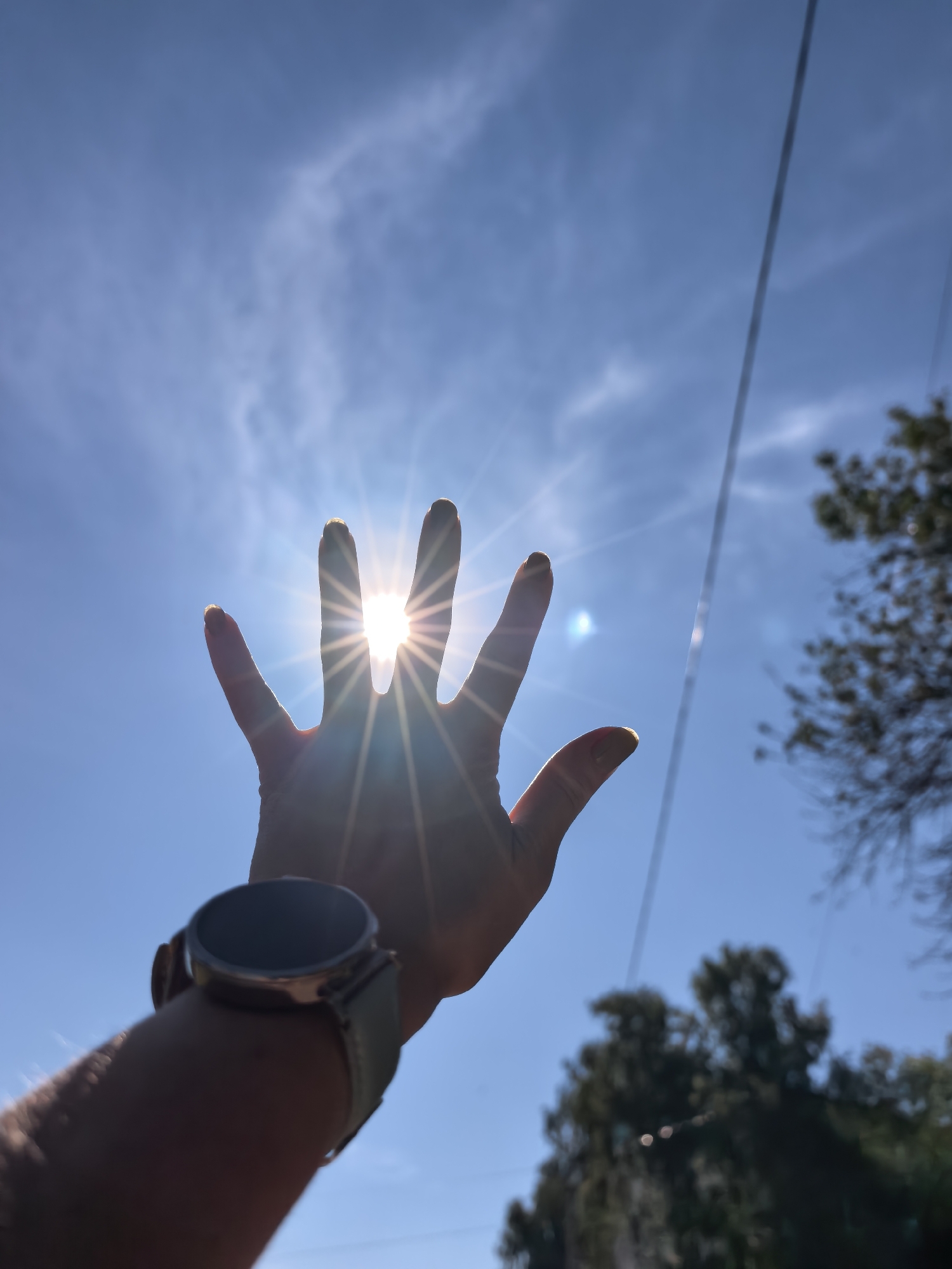 A person's hand, silhouetted against a bright blue sky with wispy clouds, is held up with fingers spread apart, allowing the sun to shine through the gaps. The sun's rays create a starburst effect. A smartwatch with a light-colored band is visible on the person's wrist. In the background, out of focus, are the silhouettes of trees and a power line. The scene appears to be outdoors, during the daytime, with bright sunlight. There is no information in the image to determine the location in the city of Sterlitamak, Russia.