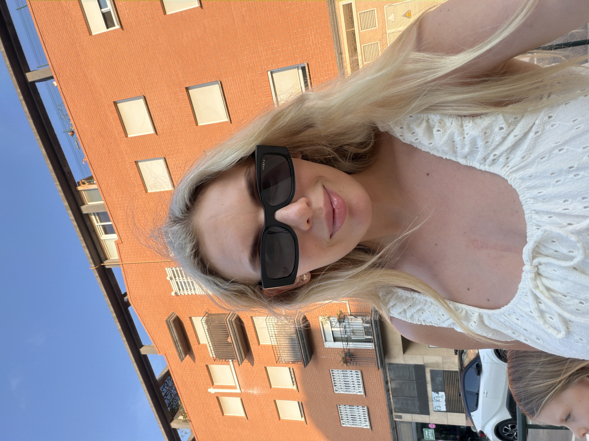 The media file is a portrait of a young woman outdoors, with a brick building and clear blue sky in the background. The woman has long blonde hair and is wearing sunglasses and a white top with eyelet details. She appears to be taking a selfie. The scene suggests a sunny day, likely during daylight hours. The building in the background is a multi-story brick structure with several windows and balconies. A white car is visible in the lower right portion of the image. The overall mood is casual and relaxed. The sunglasses have "Vogue" written on them.