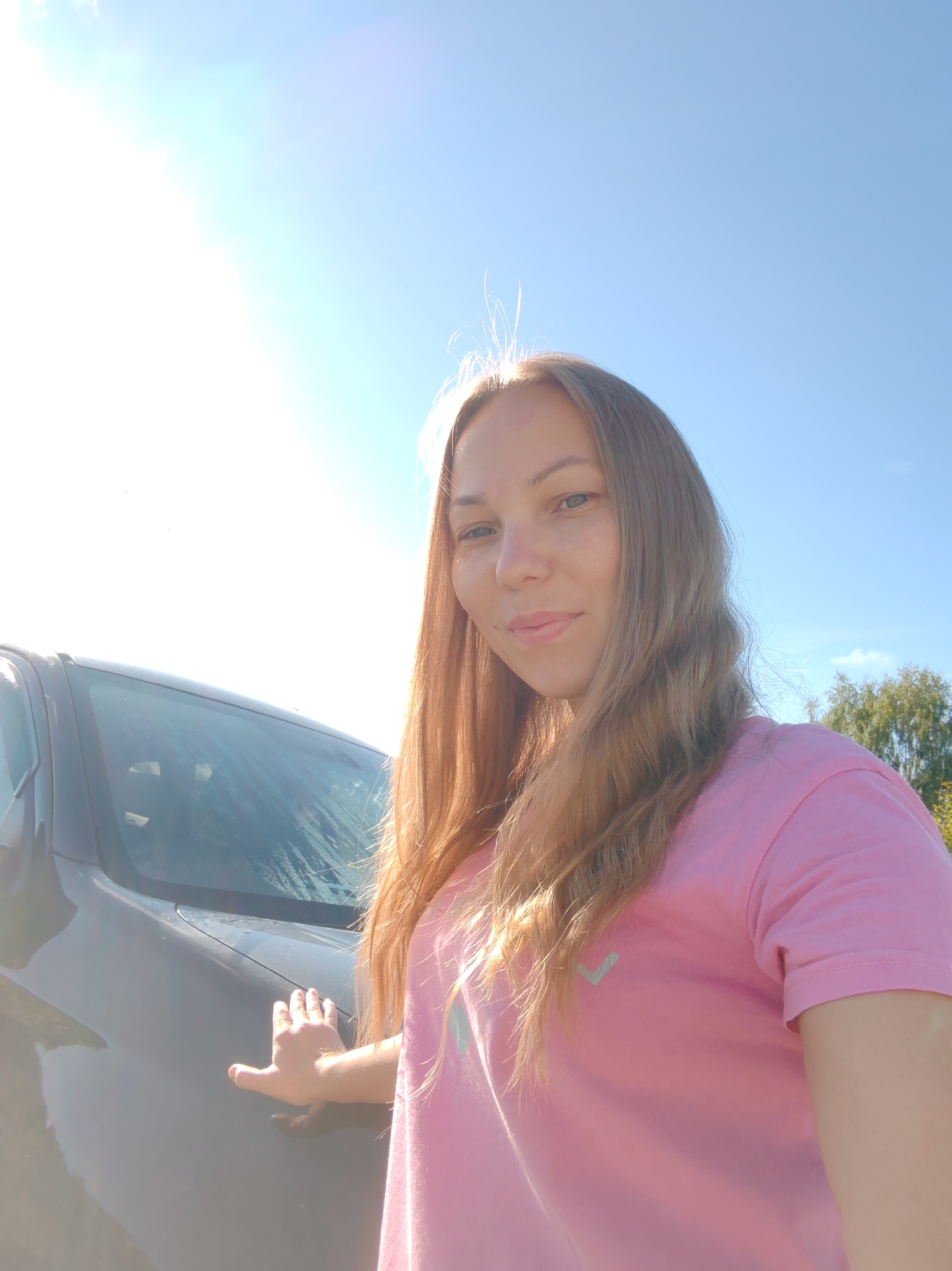 In Чухлэм, Russia, a woman with long, light brown hair and fair skin is captured in a selfie. She is wearing a pink t-shirt with a light gray checkmark logo on the left chest. The woman is standing next to a dark gray car, with her left hand resting on its front fender. The car's windshield appears wet. Behind her, the sky is a clear blue with the sun shining brightly, creating a lens flare effect in the upper left portion of the image. Trees are visible in the background on the right side. The scene suggests an outdoor setting on a sunny day.