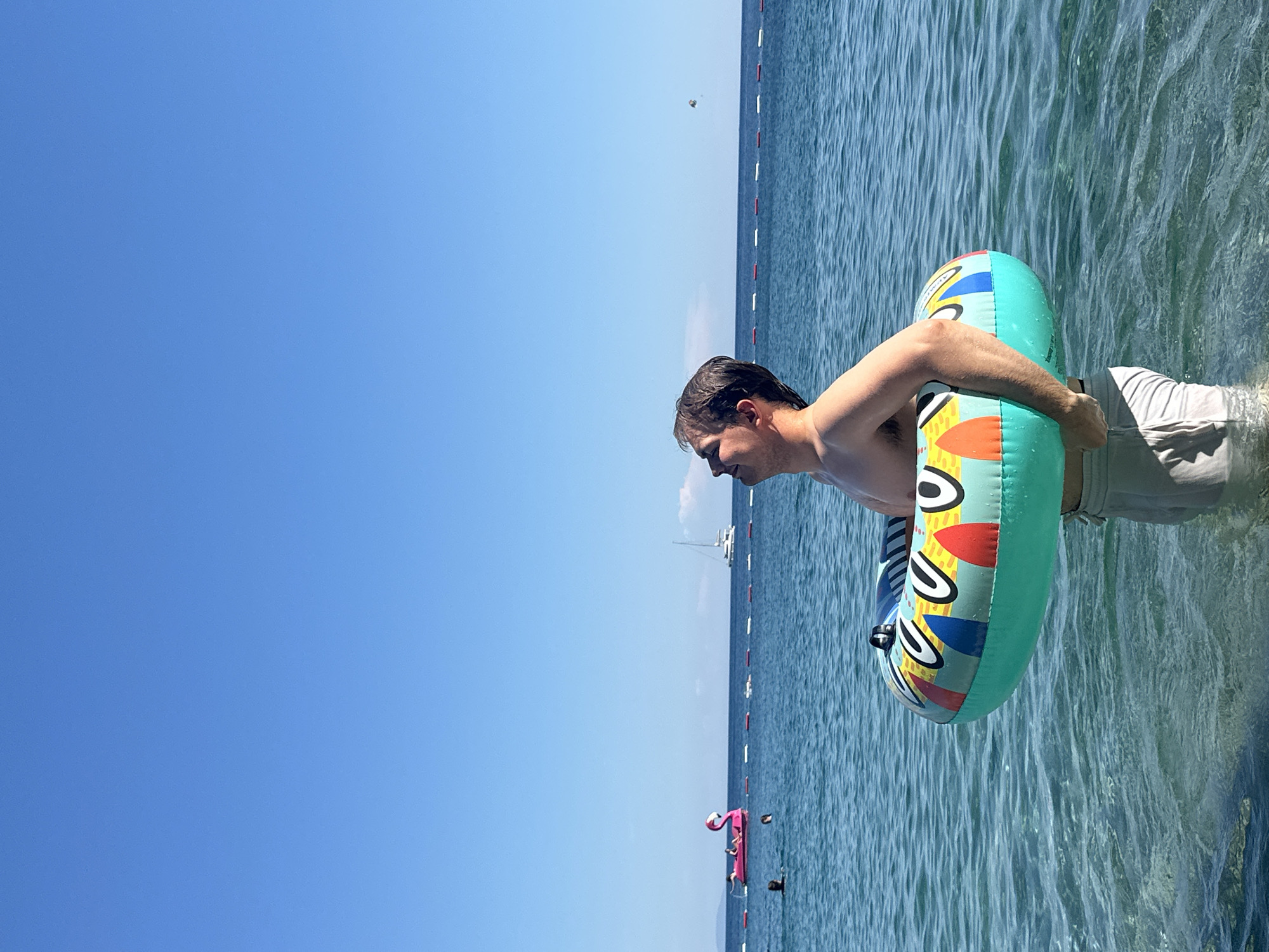 A man stands in the sea wearing swimming trunks and holding a colorful inflatable ring. In the distance, a safety line is visible in the water. Further out, there are boats and other people floating on inflatable objects, one of which is shaped like a pink flamingo. The sky is blue and clear. The location is Budva, Montenegro.