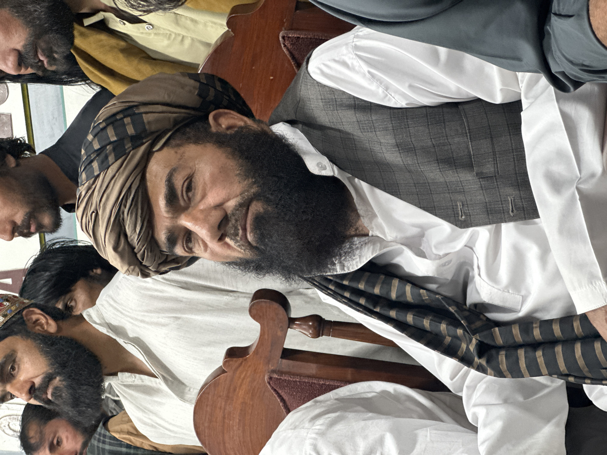 In Quetta City Tehsil, Pakistan, a man with a full beard and a patterned turban sits in a wooden chair with a burgundy cushion. He wears a white collared shirt under a gray plaid vest. Part of a horizontally-striped scarf is visible. Other people are partially visible in the background.