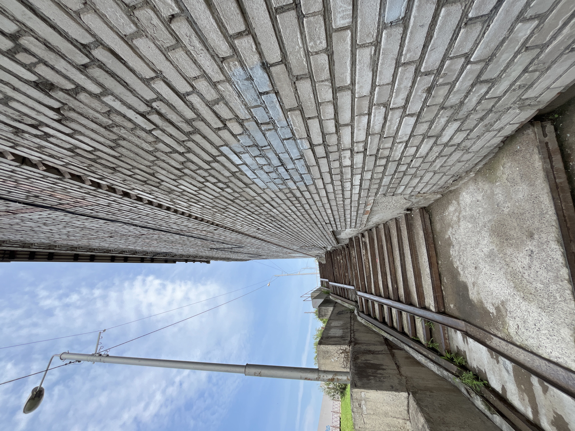 The image depicts an outdoor scene with a textured brick wall on the right side, leading upwards and away from the viewer. The wall is constructed of light-colored bricks with dark mortar lines, and a section of the wall shows faint blue paint marks. To the left of the brick wall, a concrete staircase ascends, with metal railings along its sides. Adjacent to the stairs, a concrete ramp with metal runners is visible. The ground in front of the stairs and ramp is wet concrete. A tall, grey lamppost stands on the left side of the image, with wires extending from it. In the background, a clear blue sky with scattered white clouds is present. The overall setting suggests an urban environment, likely in Krasnoyarsk, Russia, given the context. There are no people or animals visible in the image. The scene appears to be a utility area or an access point to a higher elevation within the city.