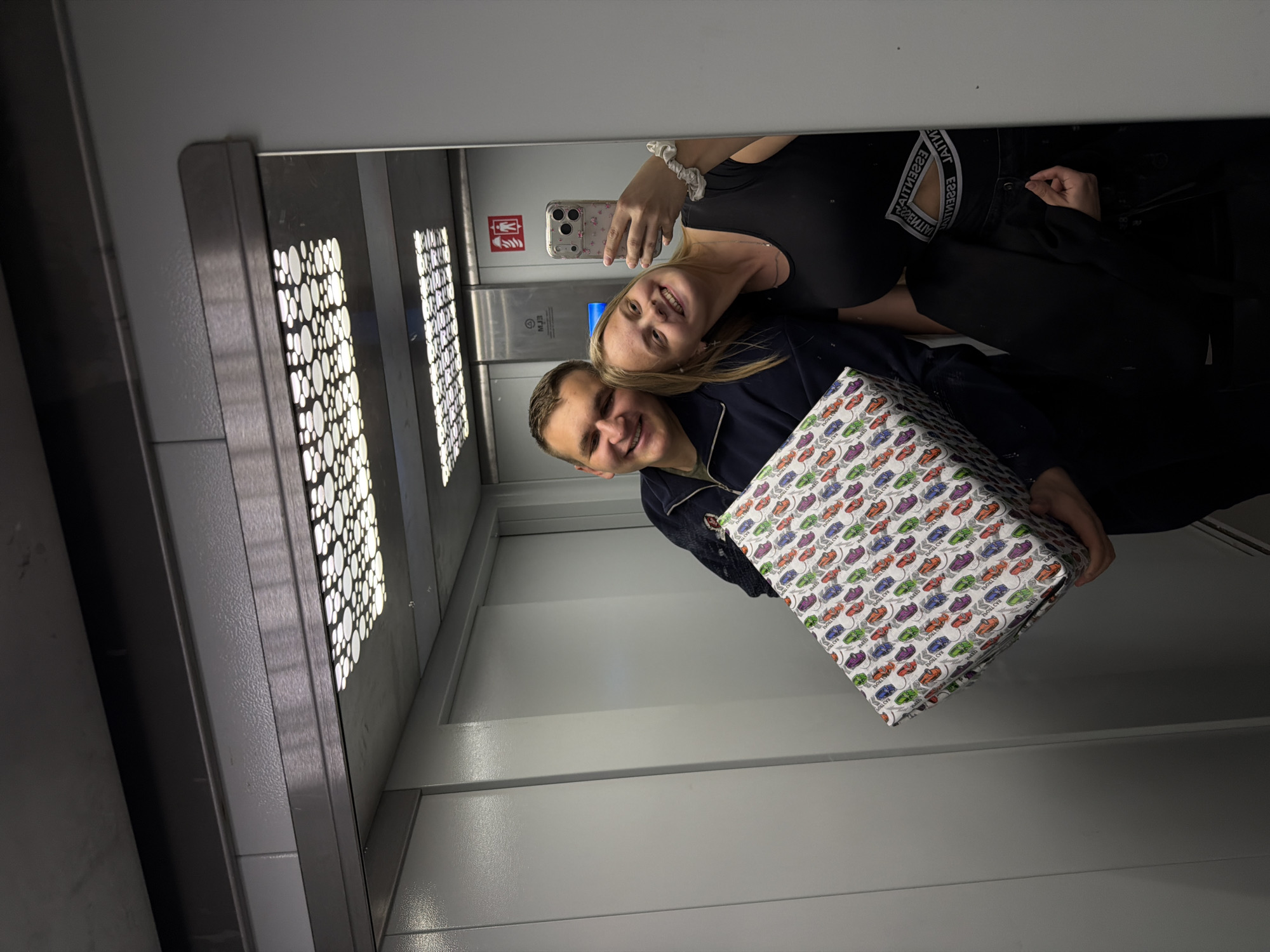 The image shows a couple taking a selfie in a mirror inside an elevator. The elevator has a patterned light fixture above and gray metal doors. The woman is holding the phone and smiling, wearing a black top and a scrunchie. The man is also smiling and holding a gift wrapped in paper with a colorful sneaker print. The wall behind them is gray with an emergency pull cord and instructions. The elevator's interior is primarily gray. Given the context, it is possible the location is within the city of Izhevsk, Russia, but the image itself does not contain any visual cues or identifying characteristics that would confirm it.
