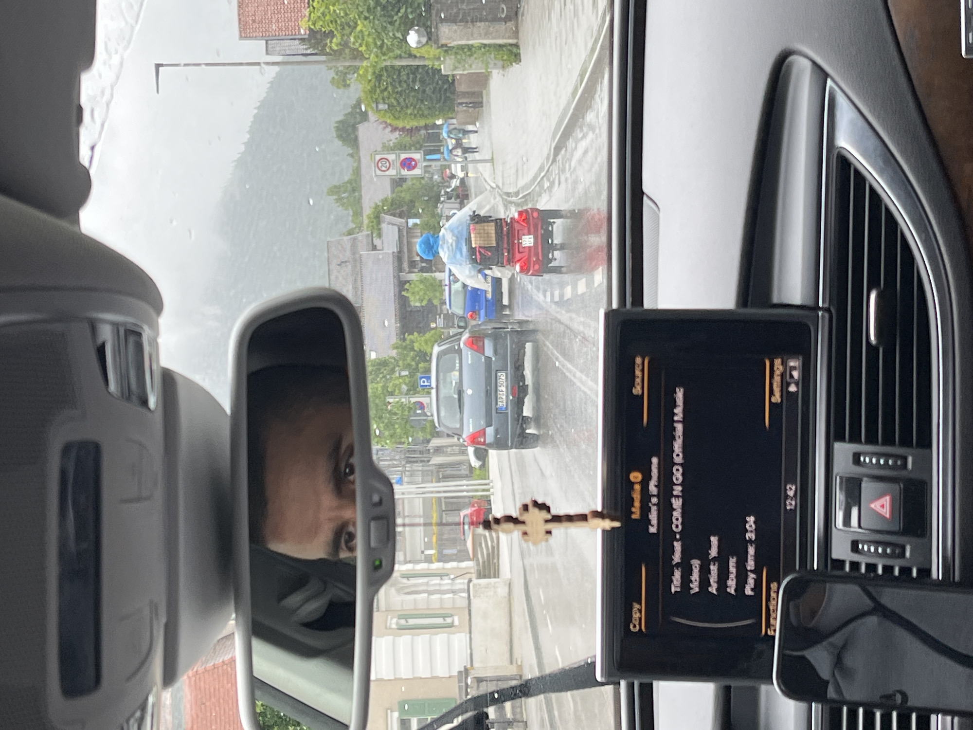 The image, taken from inside a car in Garmisch-Partenkirchen, Germany, shows a street scene on a rainy day. The car's infotainment screen displays information about the song "COME N GO" by Yeat. A wooden crucifix hangs from the rearview mirror. Outside, cars drive on a wet road. A person in a mobility scooter wears a blue rain poncho. Buildings with tiled roofs and trees are visible, and a mountain is in the background. A speed limit sign is posted.