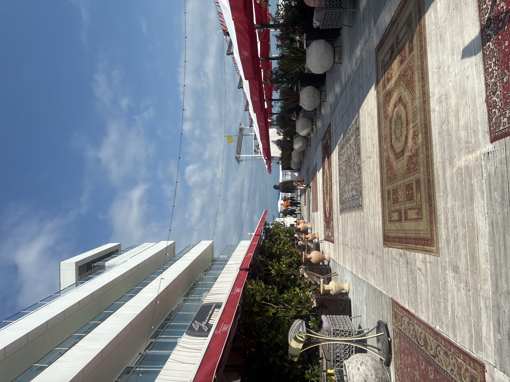 The image depicts an outdoor seating area with an open-air restaurant in Sochi, Russia. A multi-story white building with balconies and glass railings forms the backdrop on the left. In front of the building, red awnings cover outdoor seating. The ground is made of light-colored wooden planks, upon which several ornate Persian-style rugs are laid out. Large potted plants in concrete planters are placed along the walkway. Several people are walking on the pathway, some away from the camera and others towards it. The sky above is blue with scattered white clouds. The scene suggests a sunny day at a seaside resort.