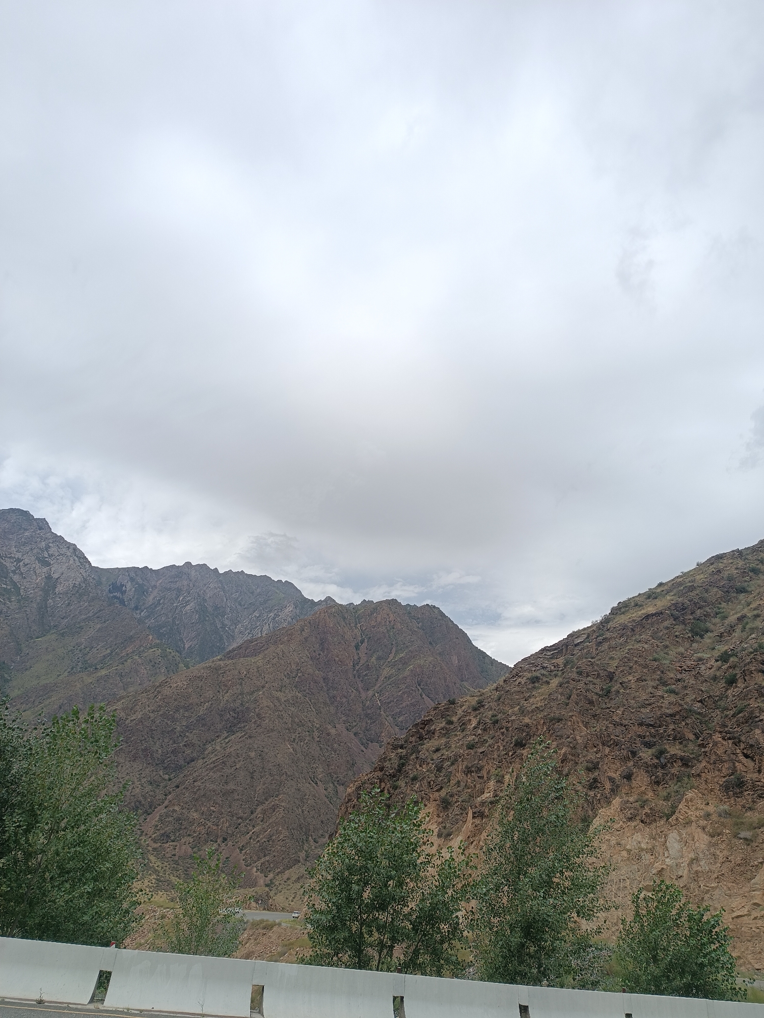The image depicts a cloudy day in the mountainous terrain surrounding Sasyk-Bulak, Kyrgyzstan. In the foreground, a white concrete barrier runs horizontally across the lower portion of the frame, with glimpses of vegetation and a road visible beyond it. The mid-ground is dominated by rugged, rocky mountains in shades of brown and green, with some sparse vegetation clinging to the slopes. In the distance, more distant mountain ranges are visible, fading into the atmospheric haze. The sky is overcast with thick, gray clouds. A few vehicles are discernable on a winding road in the middle distance, suggesting the presence of a route through the mountains. The overall impression is one of a natural, somewhat desolate landscape under a somber sky.