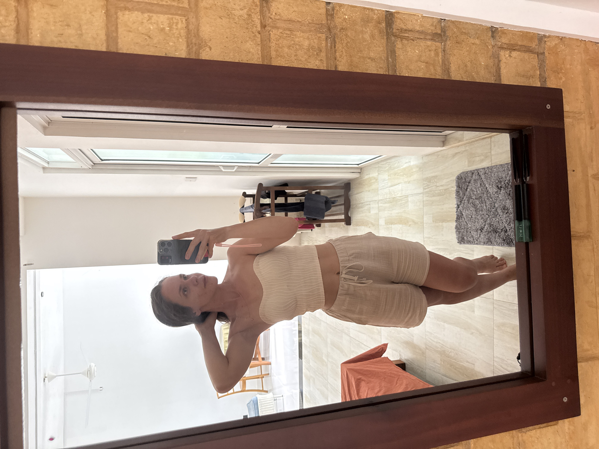 The image shows a woman taking a mirror selfie inside a building with tan brick walls in Thalaramba, Sri Lanka. She's holding a smartphone with a dark case in her right hand. The woman is wearing a cream-colored, strapless top and matching shorts. Her hair is dark and pulled back, and she is looking at her reflection in the mirror.
The mirror has a dark brown wooden frame. The reflection shows a room with light-colored tiled flooring. Visible in the reflection are a ceiling fan, a clothing rack, a gray rug, a light-colored chair, a blue object, and an orange cloth. The reflection also shows a window.