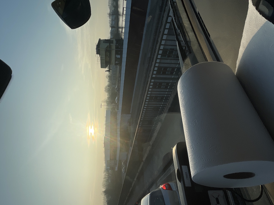 The image depicts a panoramic view from the interior of a vehicle, looking through its windshield during either sunrise or sunset, indicated by the low sun casting warm light and lens flare across the scene. A large, white roll of paper towels is positioned prominently on the vehicle's dashboard in the foreground, partially unrolled onto a dark surface.
In the midground, a multi-lane road or bridge is visible, with several passenger cars present. A dark metal railing runs along the edge of this structure.
The background reveals a large infrastructure complex associated with the Iffezheim Lock (Schleuse Iffezheim) on the Rhine River in Iffezheim, Germany. To the left, a modern, multi-story white building with a flat roof and extensive glazing is discernible, possibly an administrative or customs facility. To the right, a distinct, multi-level control tower with industrial features overlooks a body of water, identifiable as the Rhine River. Beyond the structures, a line of deciduous trees forms the horizon against a clear, pale sky. No individuals are clearly identifiable.