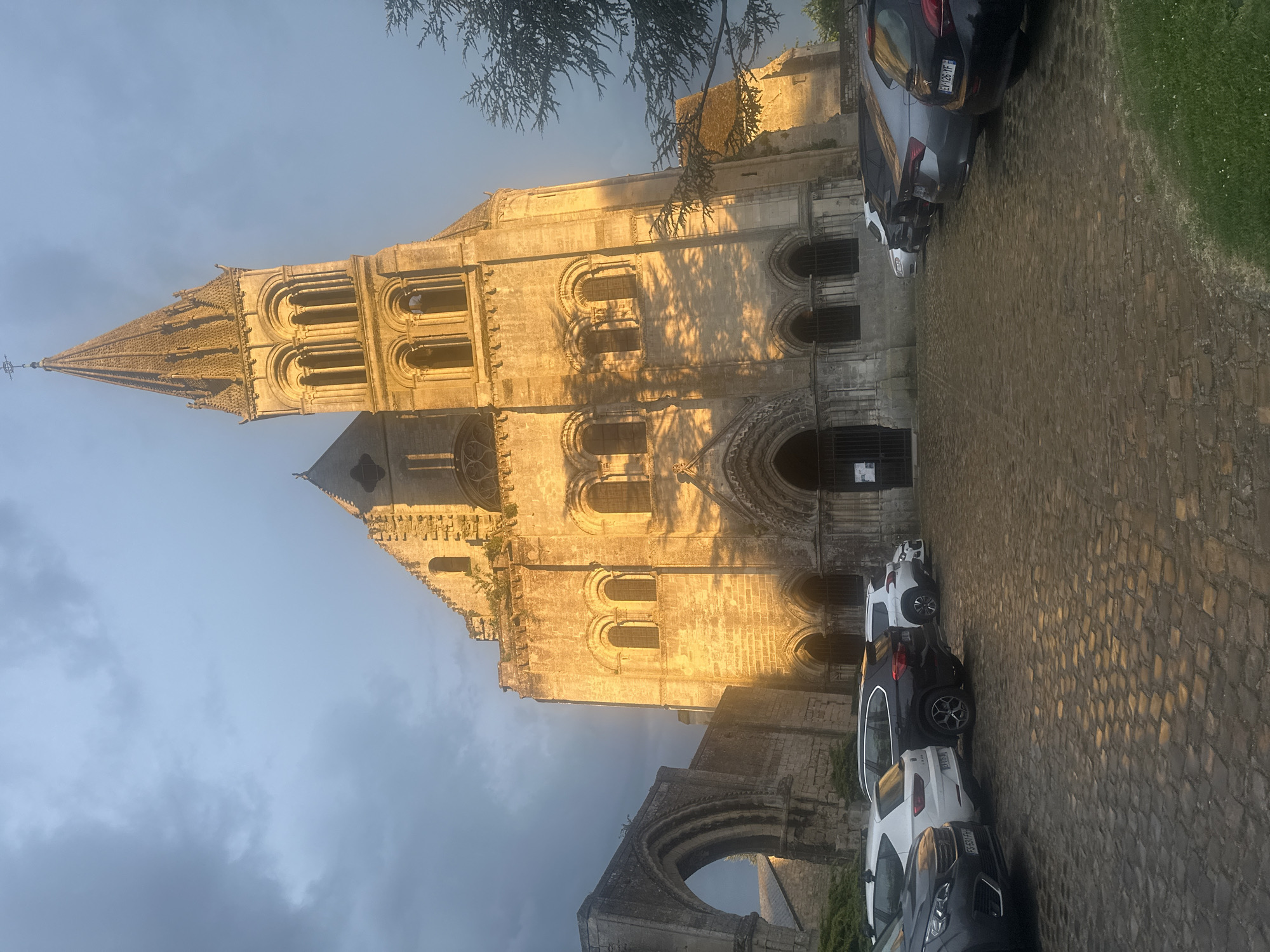 The image captures the facade of the Church of Saint-Leu-d'Esserent in Saint-Leu-d'Esserent, France. The church is constructed of light-colored stone. It is a historic building with arched windows and intricate architectural details. The church's tall spire extends towards the sky. Parked vehicles line the cobblestone road adjacent to the church. The sky above is filled with dark clouds. The image is shot from a low angle, emphasizing the height of the building.