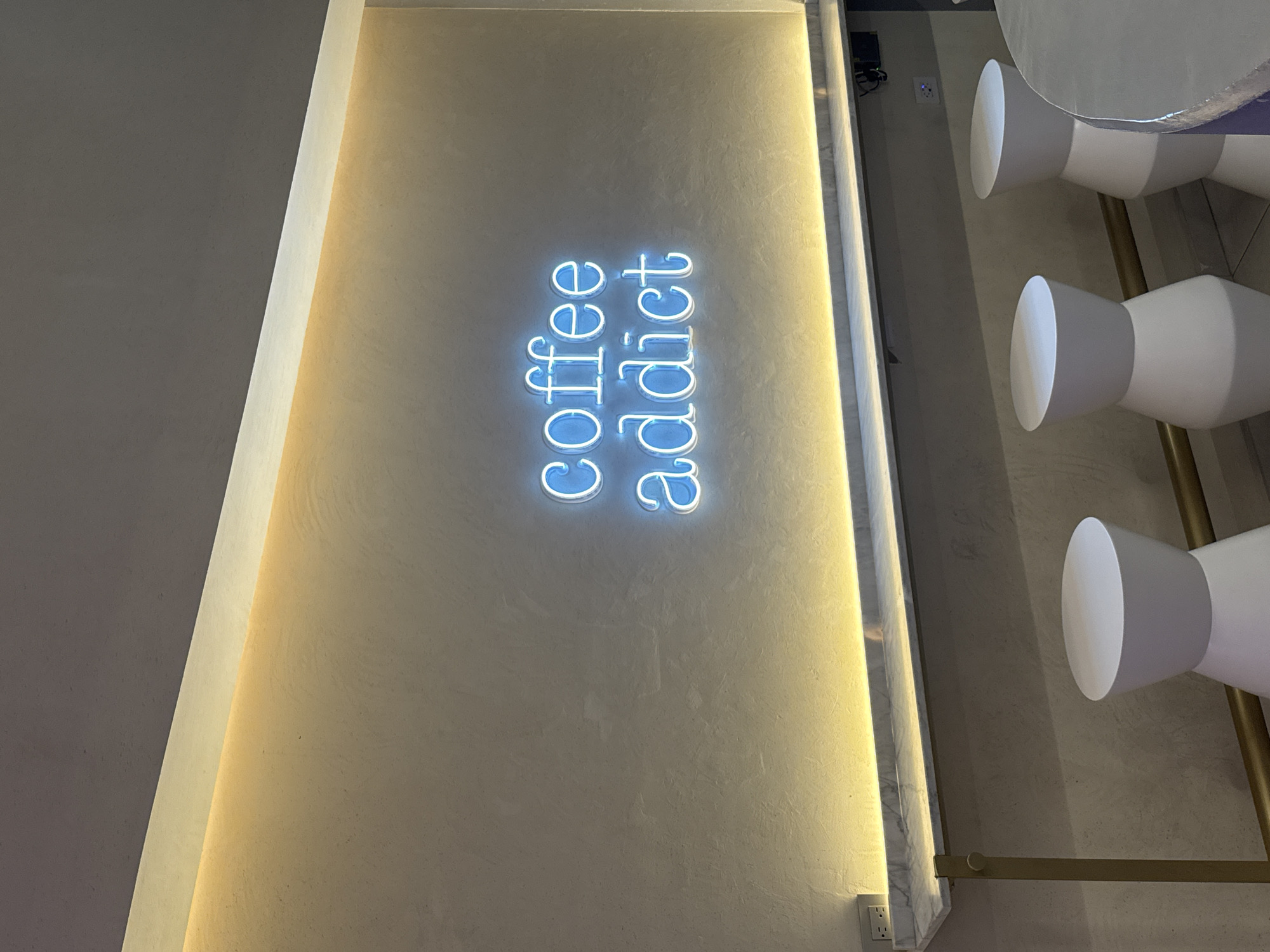 A vertical indoor shot features a sign reading "coffee addict" in blue neon lettering affixed to a textured, off-white wall. To the left, a smooth, light gray wall is separated from the sign wall by a thin strip of bright light, likely a recessed fixture. To the right, a strip of wall reveals white and light gray cylindrical light fixtures arranged in a vertical stack, mounted on a gold-colored bar. A power outlet is visible at the bottom right of the frame. The setting is likely a cafe or similar establishment. The location information given does not directly impact the description of the image.